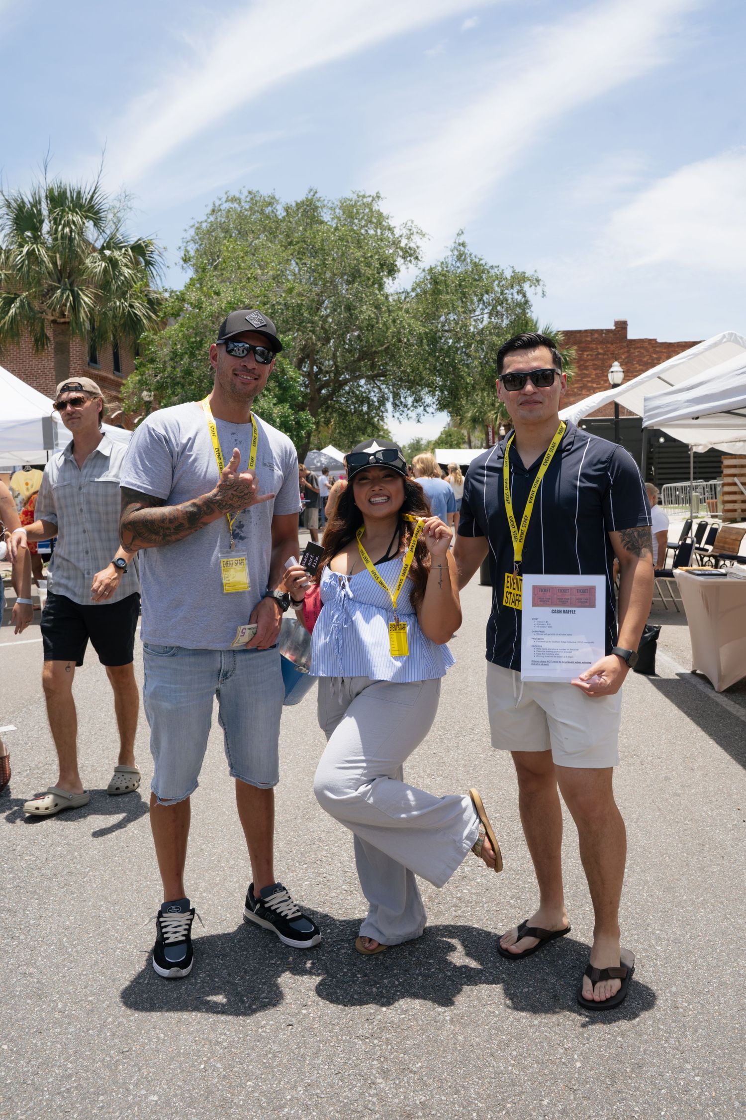 Three friends pose outdoors at a sunny festival, wearing lanyards and casual summer outfits, holding an award certificate.