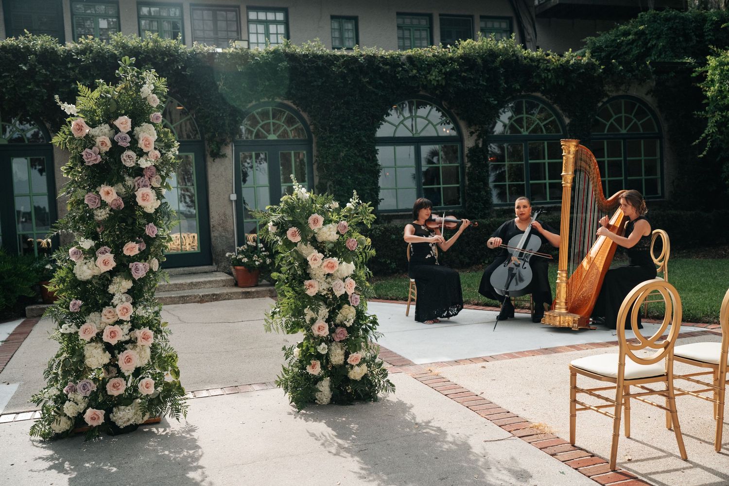 Elegant outdoor wedding ceremony with floral columns, string quartet, and harpist performing on a sunlit garden patio.