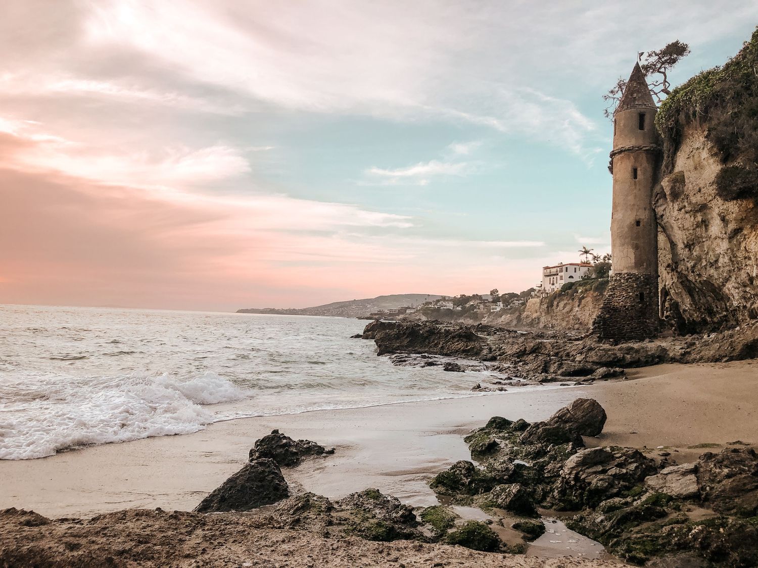 Scenic coastal beach landscape with rocky cliffs and a lighthouse tower against a pastel sunset sky.