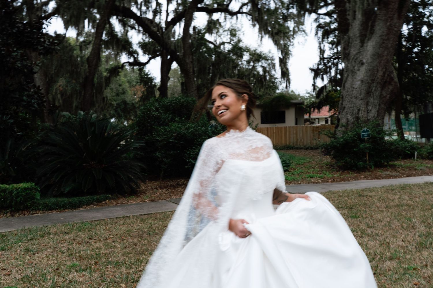 A bride in a flowing white wedding dress twirls and dances outdoors among trees and greenery.