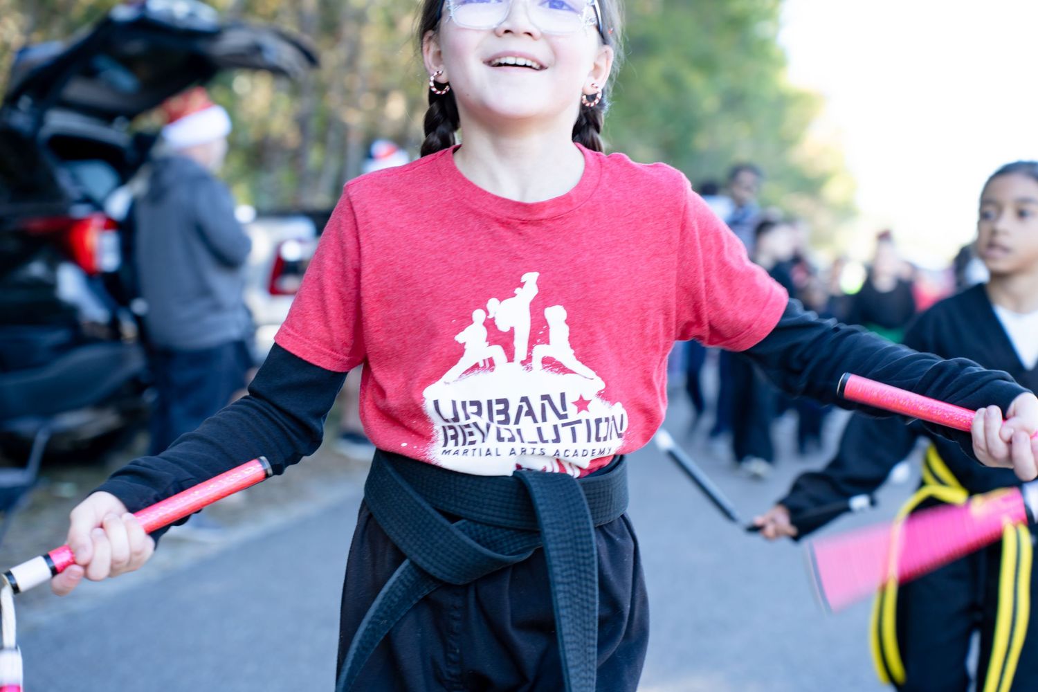 Young girl smiling in Urban Revolution red t-shirt, holding pink batons outdoors at a martial arts event.