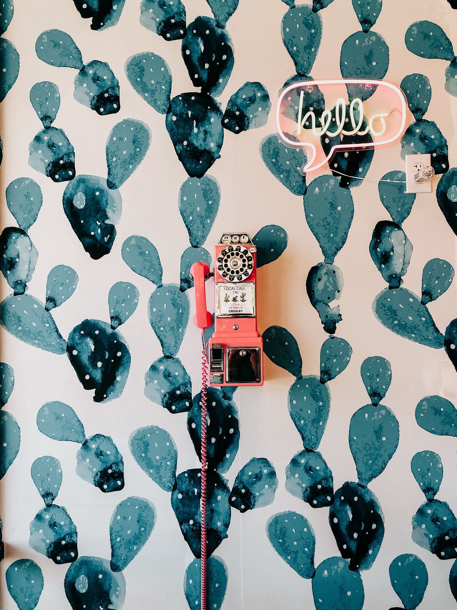 A retro pink payphone mounted on a wall with blue cactus patterned wallpaper. In San Diego, California