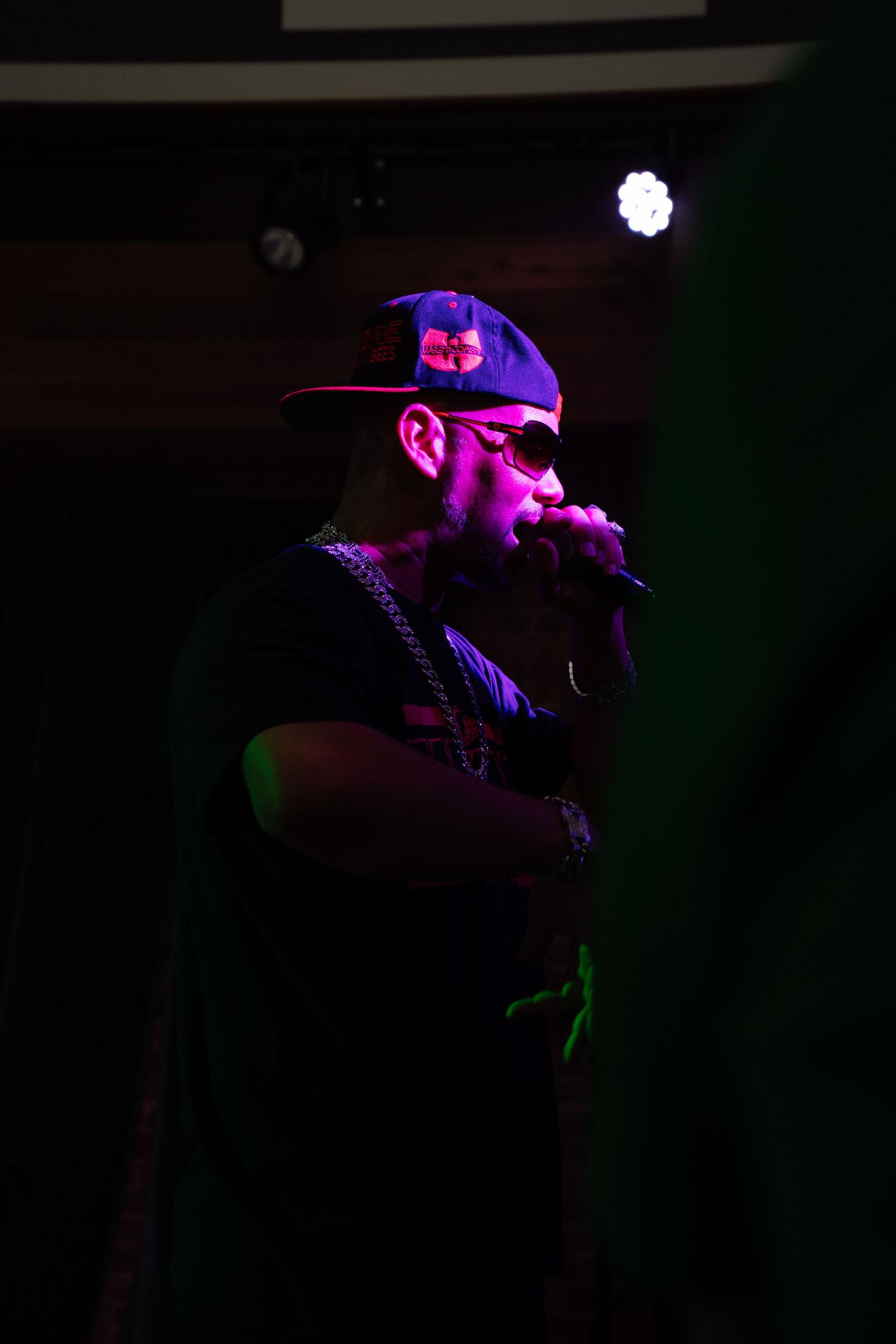 A rapper performs on stage under purple lighting, wearing a snapback cap, sunglasses, and chain necklace while holding a microphone.