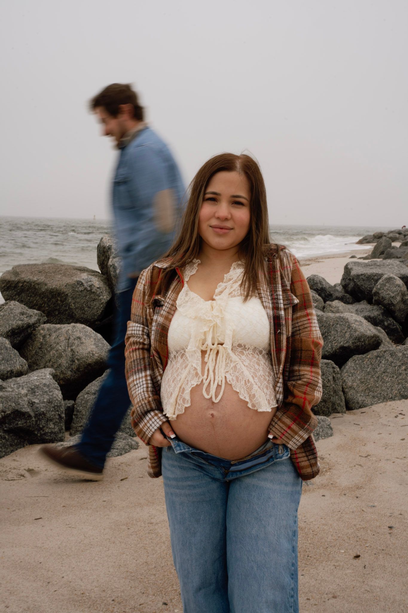 A person wearing a brown plaid flannel and white lace top poses against a rocky beach landscape.