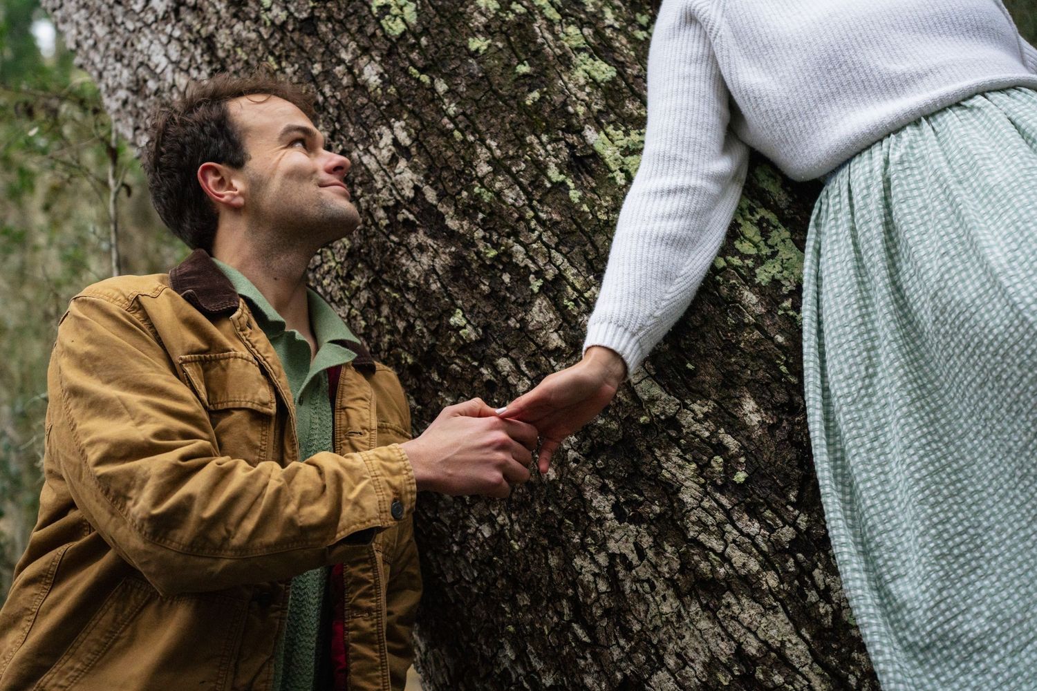 A couple in vintage clothing embraces romantically against a natural outdoor backdrop with greenery.
