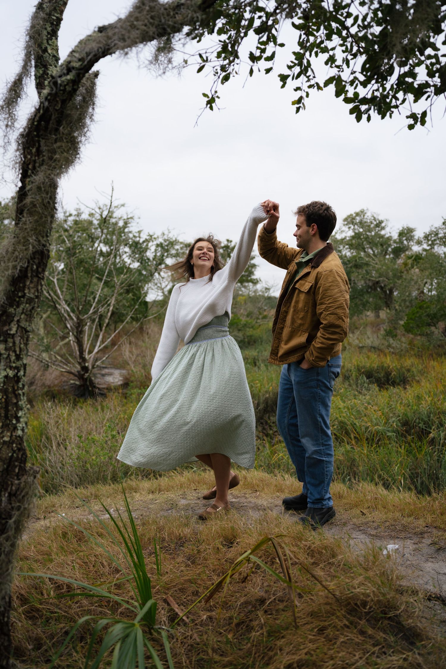 A couple in casual attire dance together in a natural outdoor setting with trees and grass in the background.