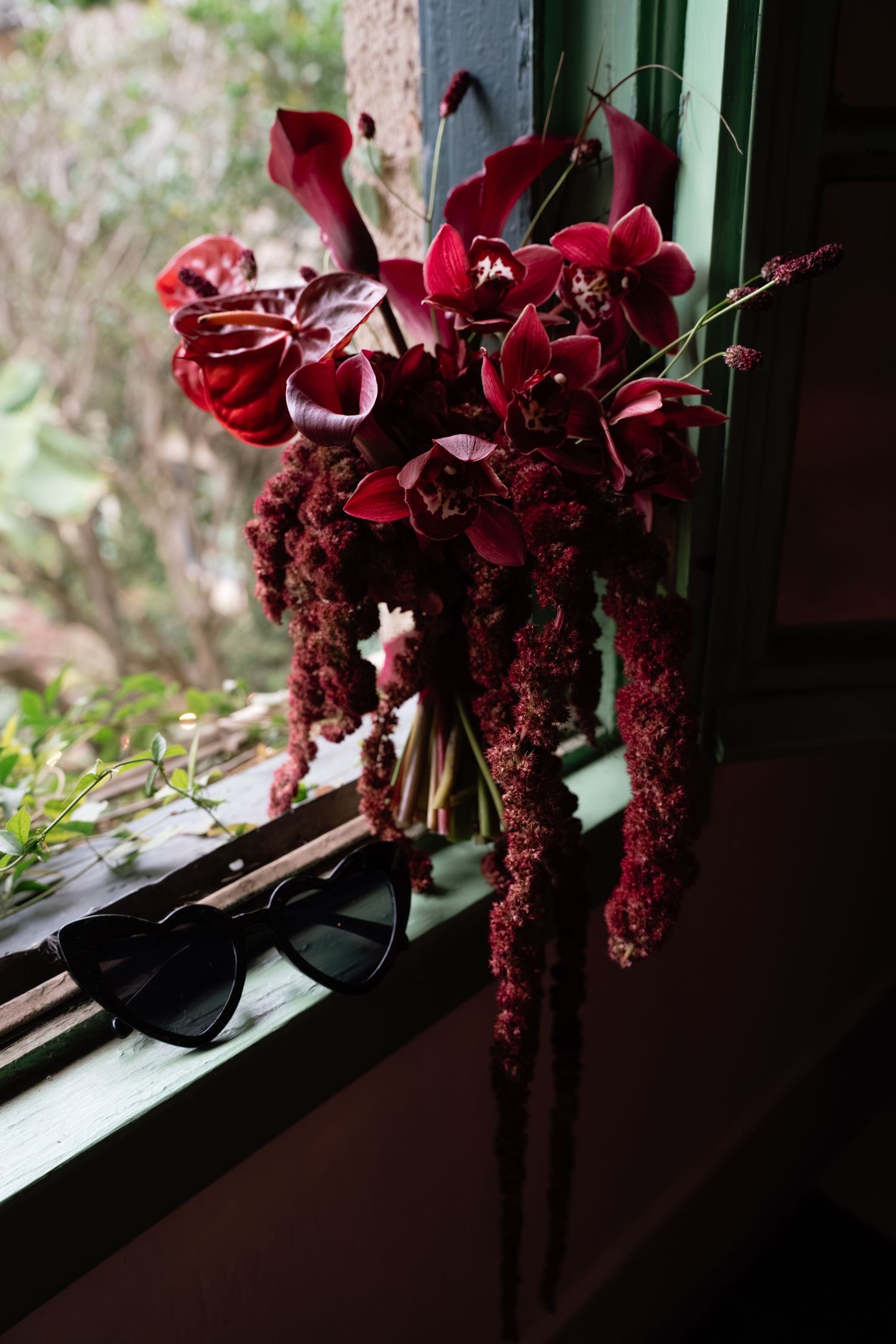 Dark red orchids in a glass vase sit on a windowsill casting dramatic shadows in natural light.