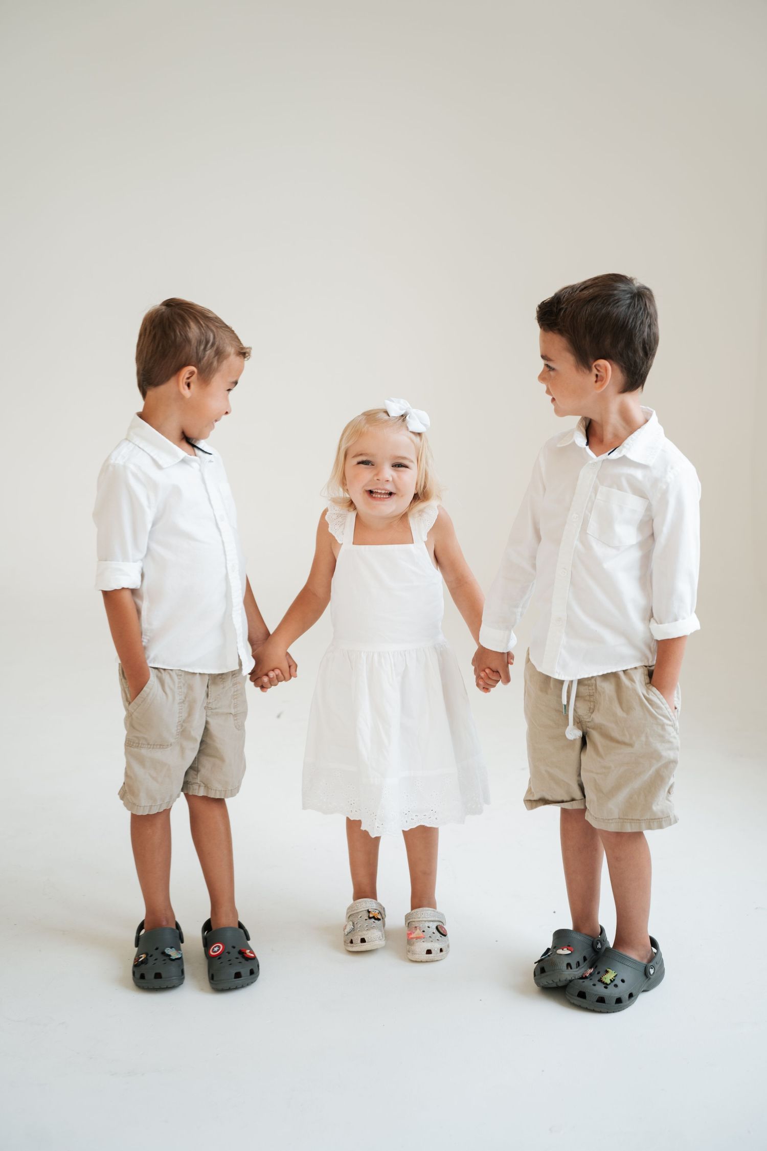Three children in coordinating white and khaki outfits stand together holding hands against a light gray background.