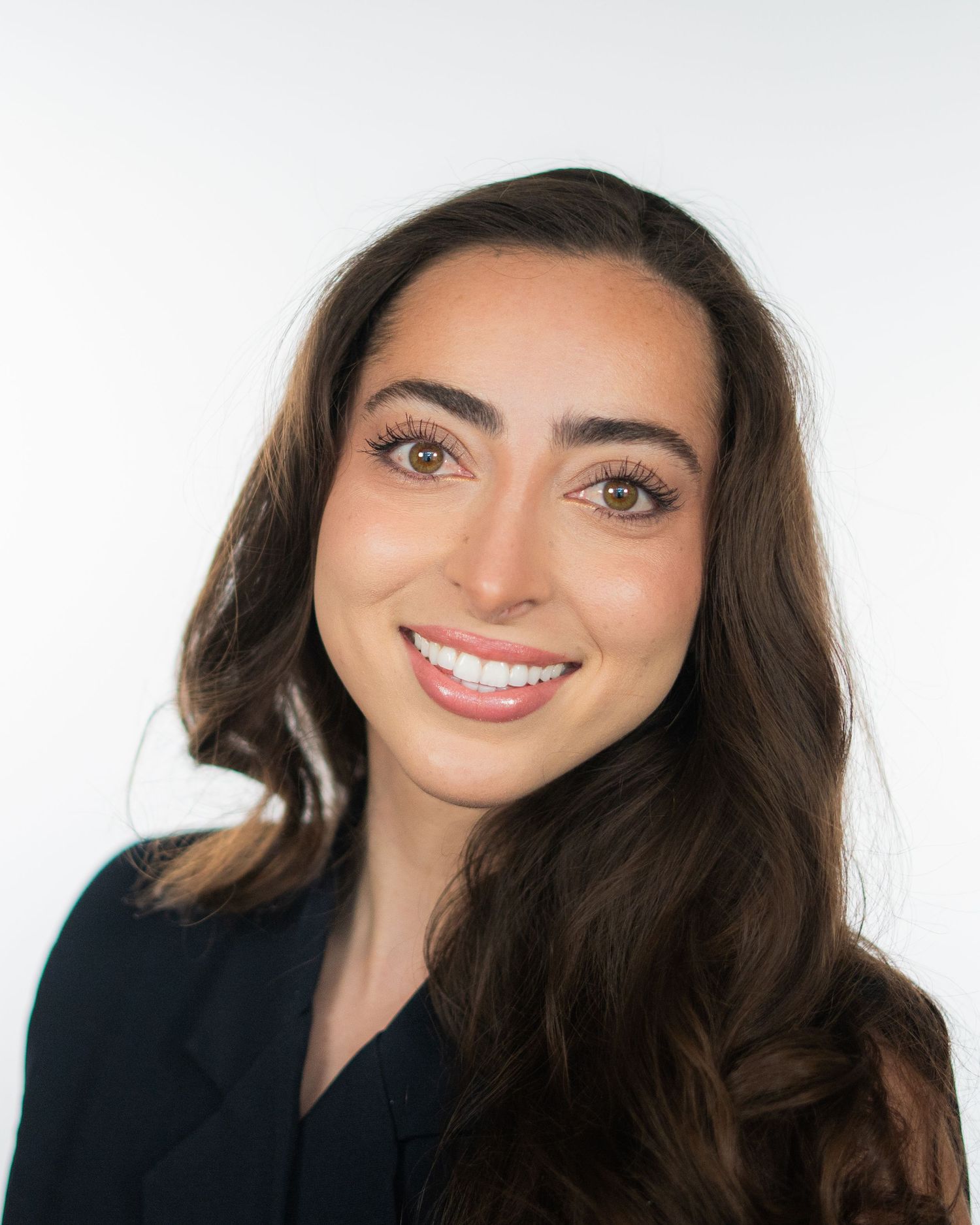 Professional headshot of a smiling young woman with long brown hair wearing a black blazer against a white background.
