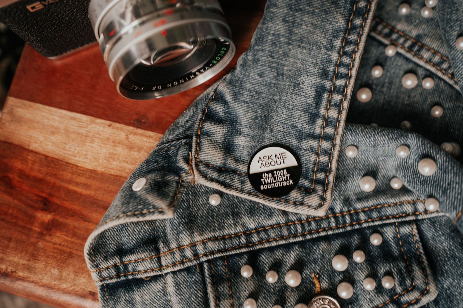 Close-up of a vintage camera lens and studded denim jacket with a pin on wooden surface.