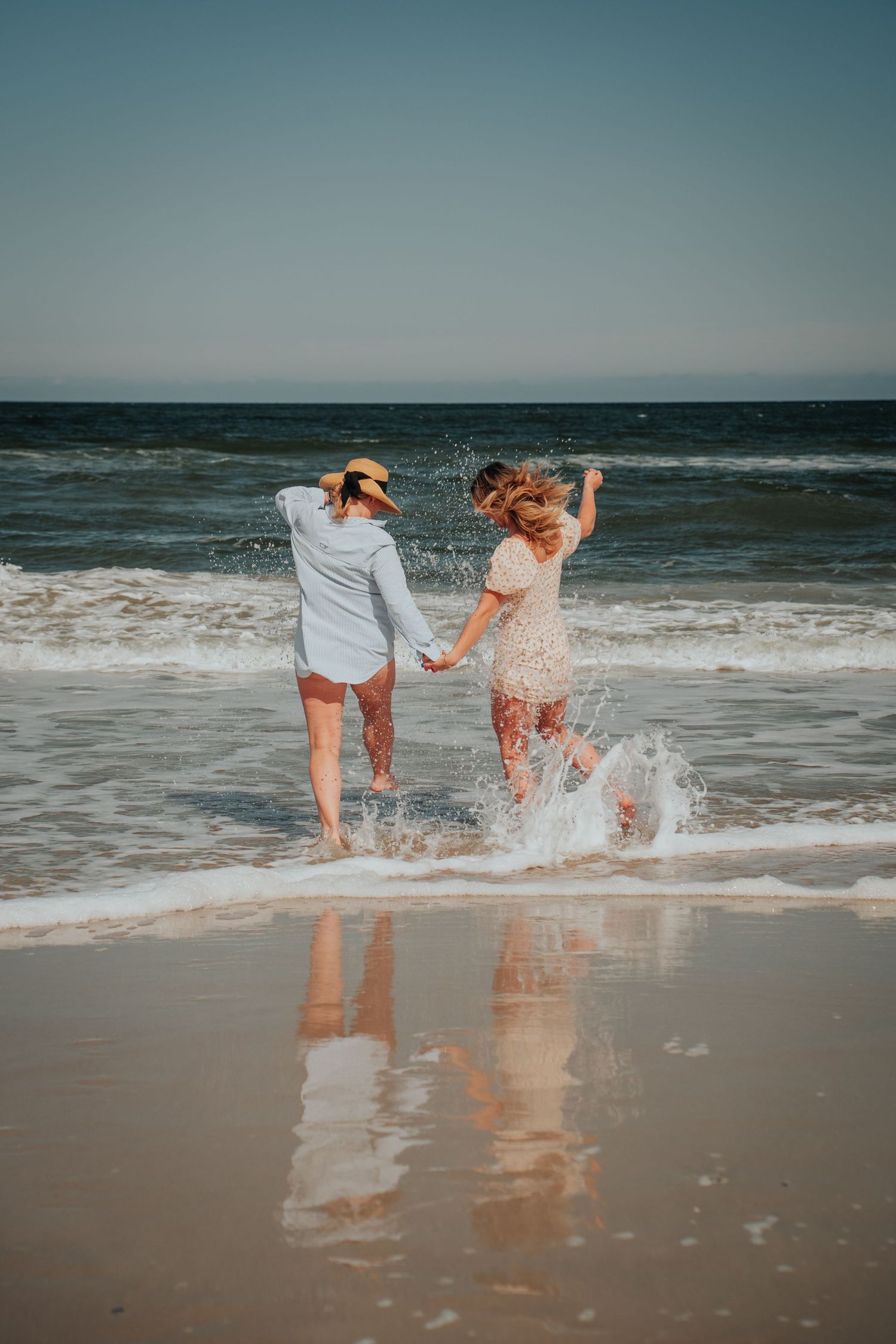 Two people holding hands and joyfully splashing in ocean waves at the beach during sunset with reflections in wet sand.