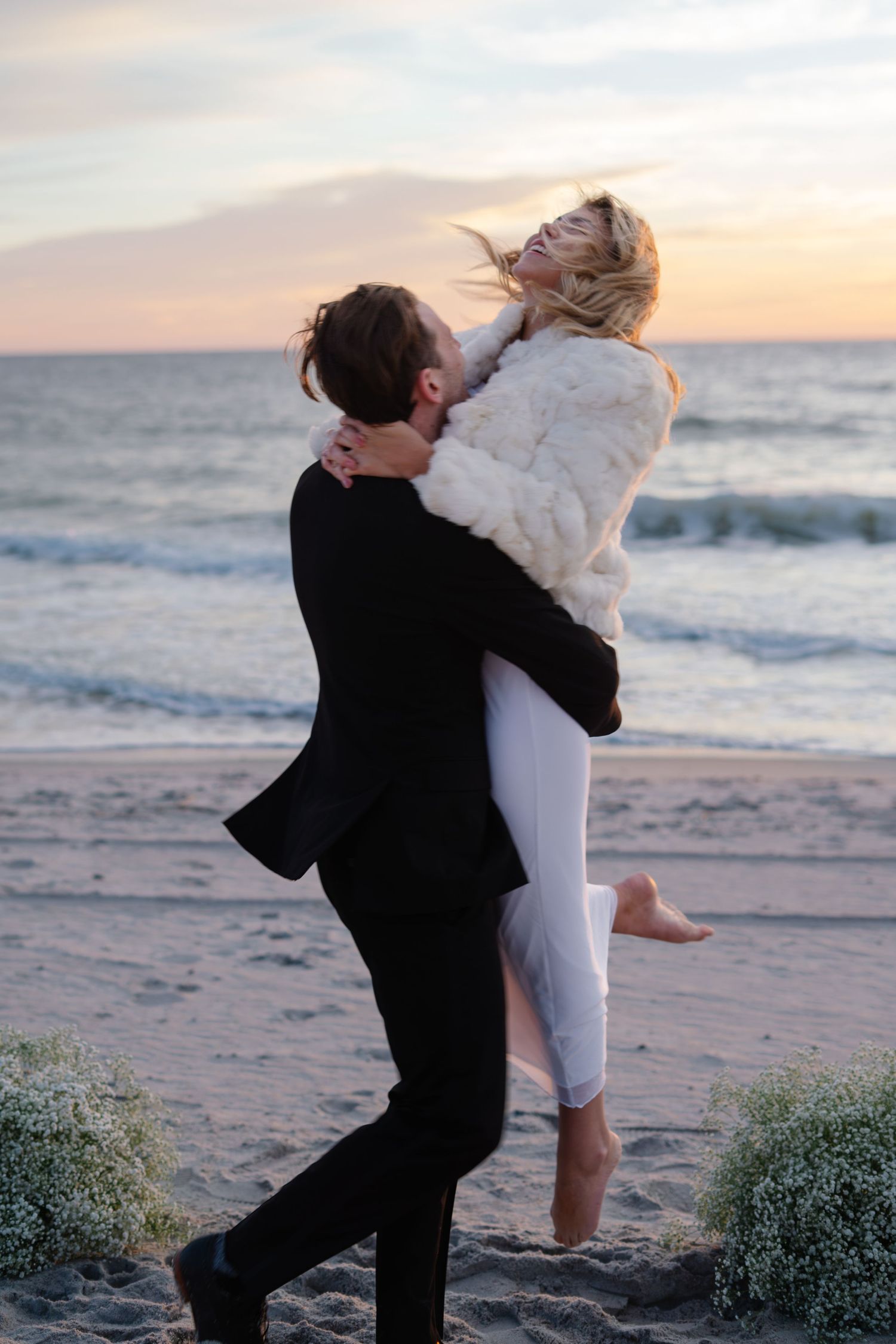 Two people share a romantic moment on the beach at sunset as waves crash on the shore in the background.