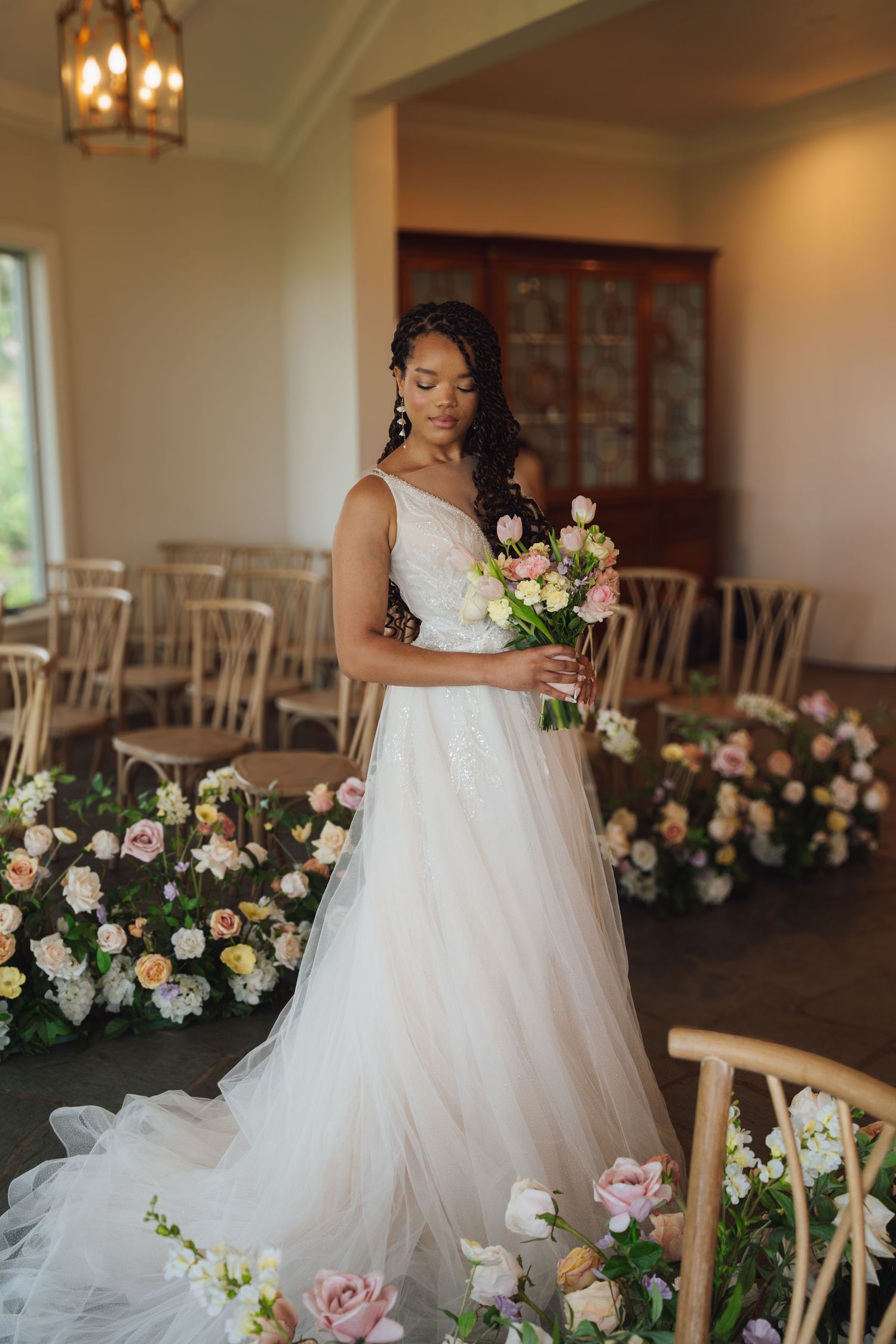 A bride in a flowing white ballgown poses elegantly beside floral arrangements at an indoor wedding venue. Azaleana Manor.
