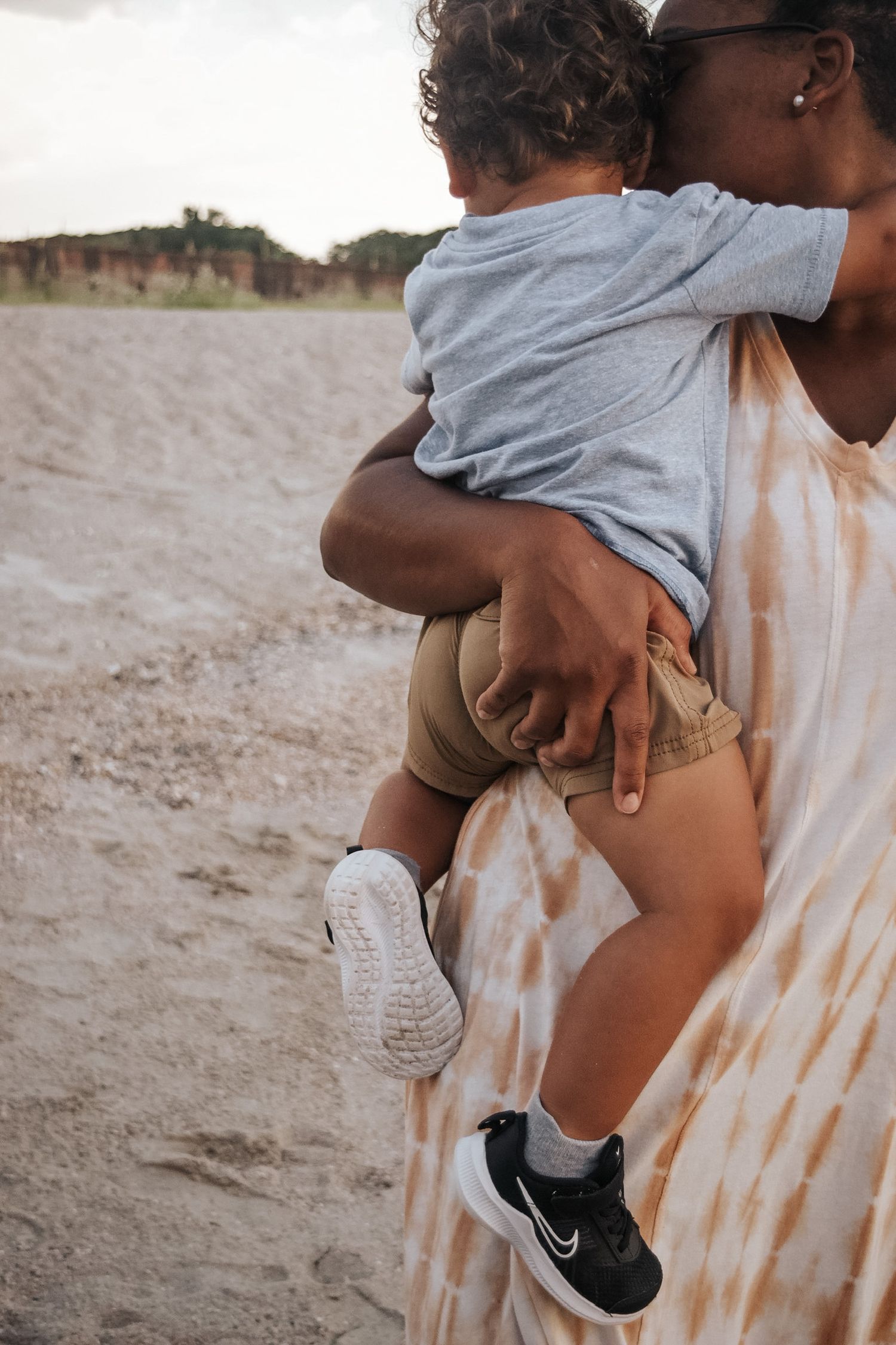Person in tie-dye dress holds small child wearing gray shirt and black sneakers on sandy beach.