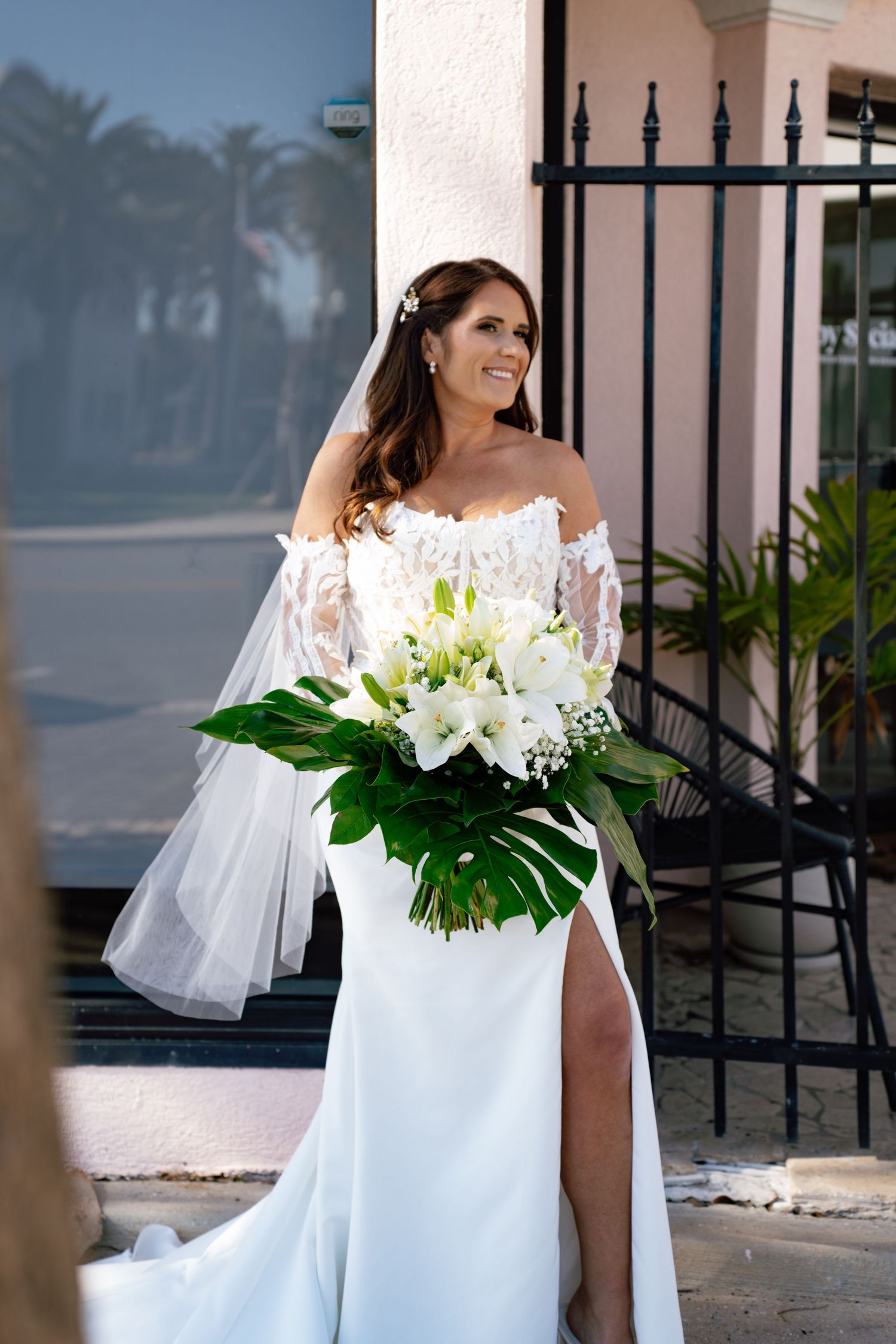 Bride in off-shoulder lace wedding gown holding white floral bouquet with tropical leaves, smiling outdoors near iron gate.