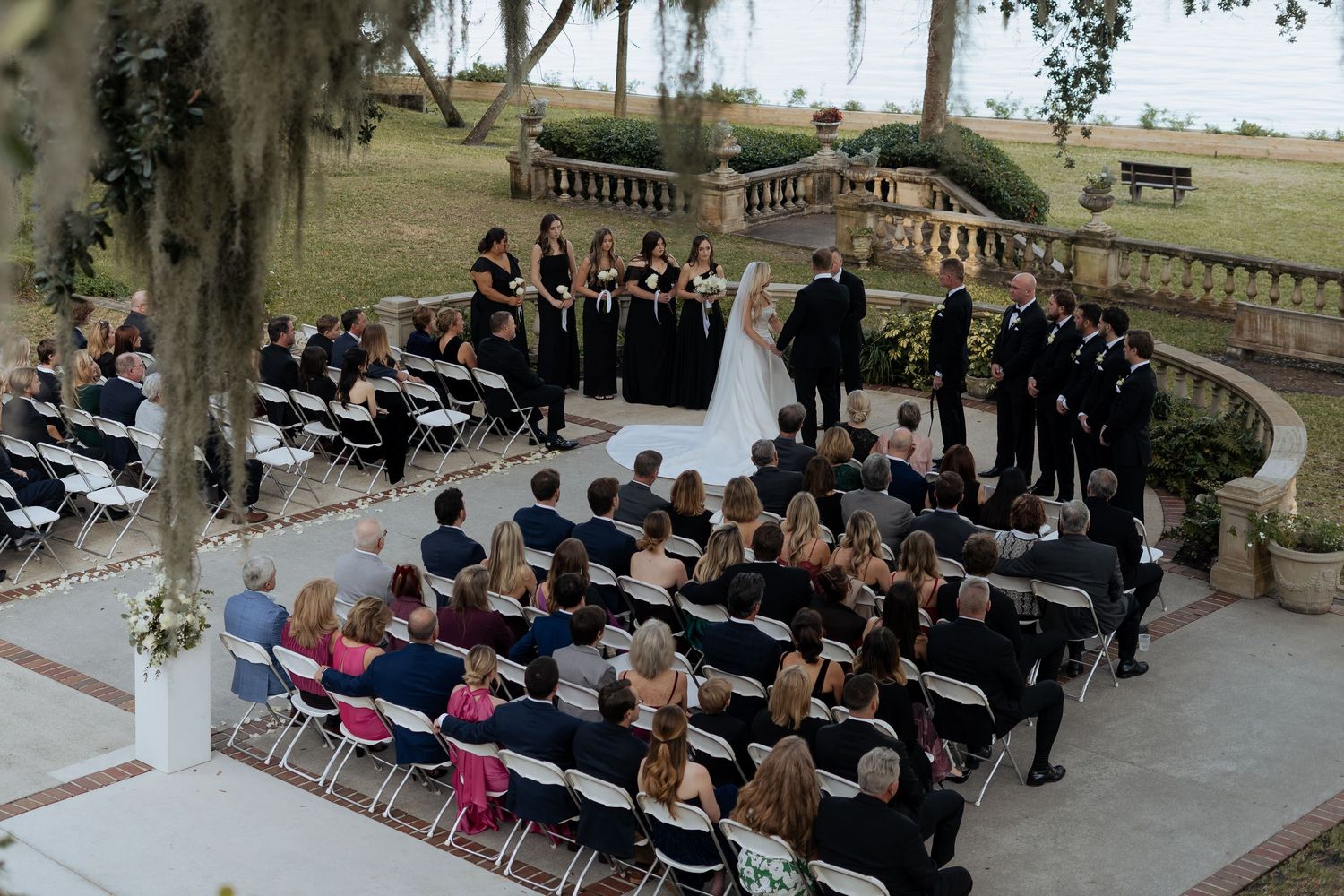 Guests seated in rows at an outdoor wedding ceremony under moss-draped oak trees in a garden setting.