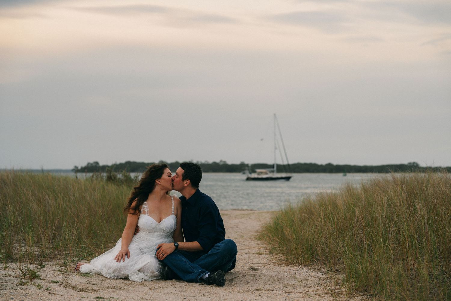 A couple sits together on a sandy beach path with tall grass and a sailboat in the distance at dusk.