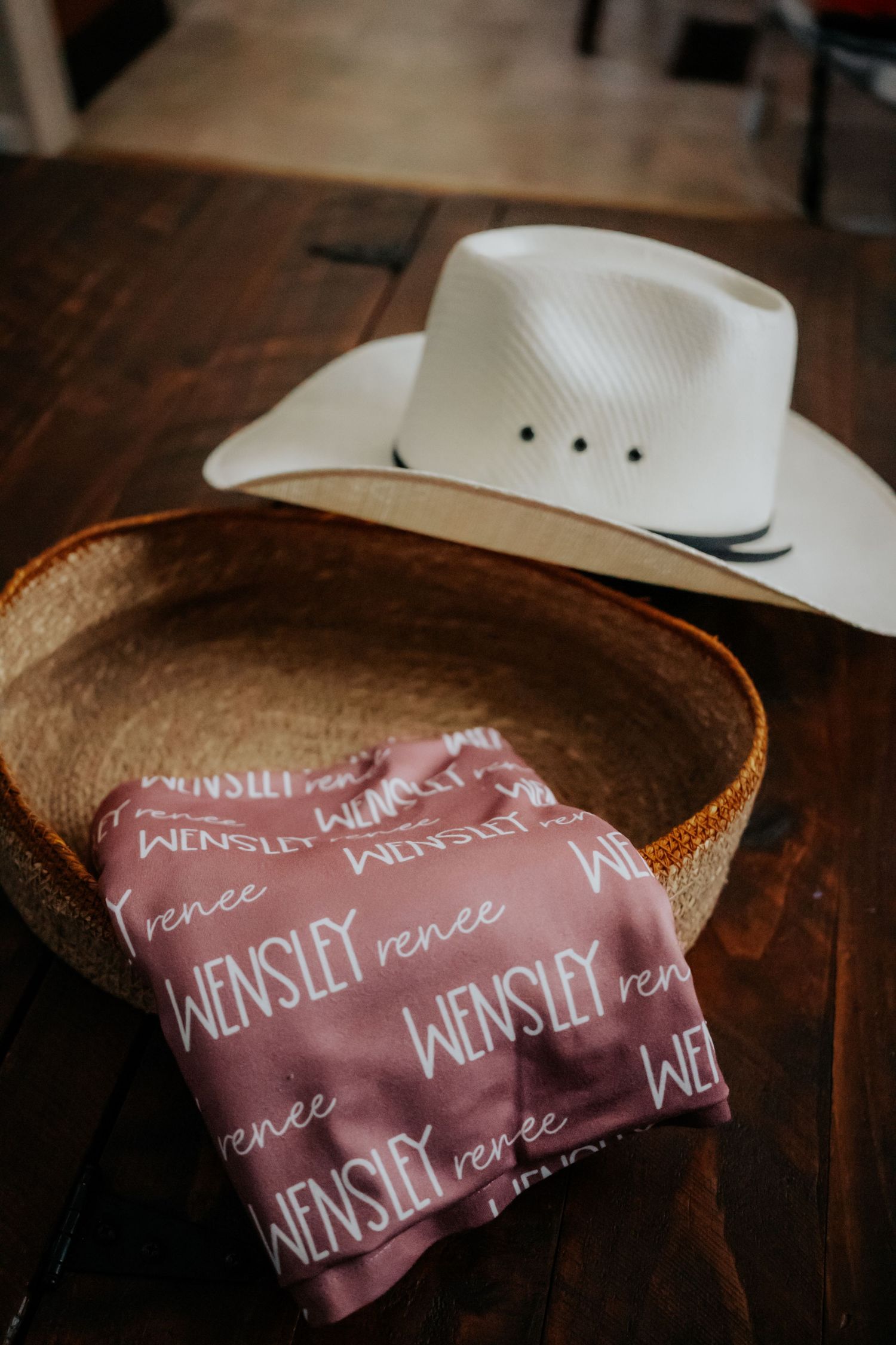 A white cowboy hat sits next to a pink personalized bandana with repeating text on a wooden table.