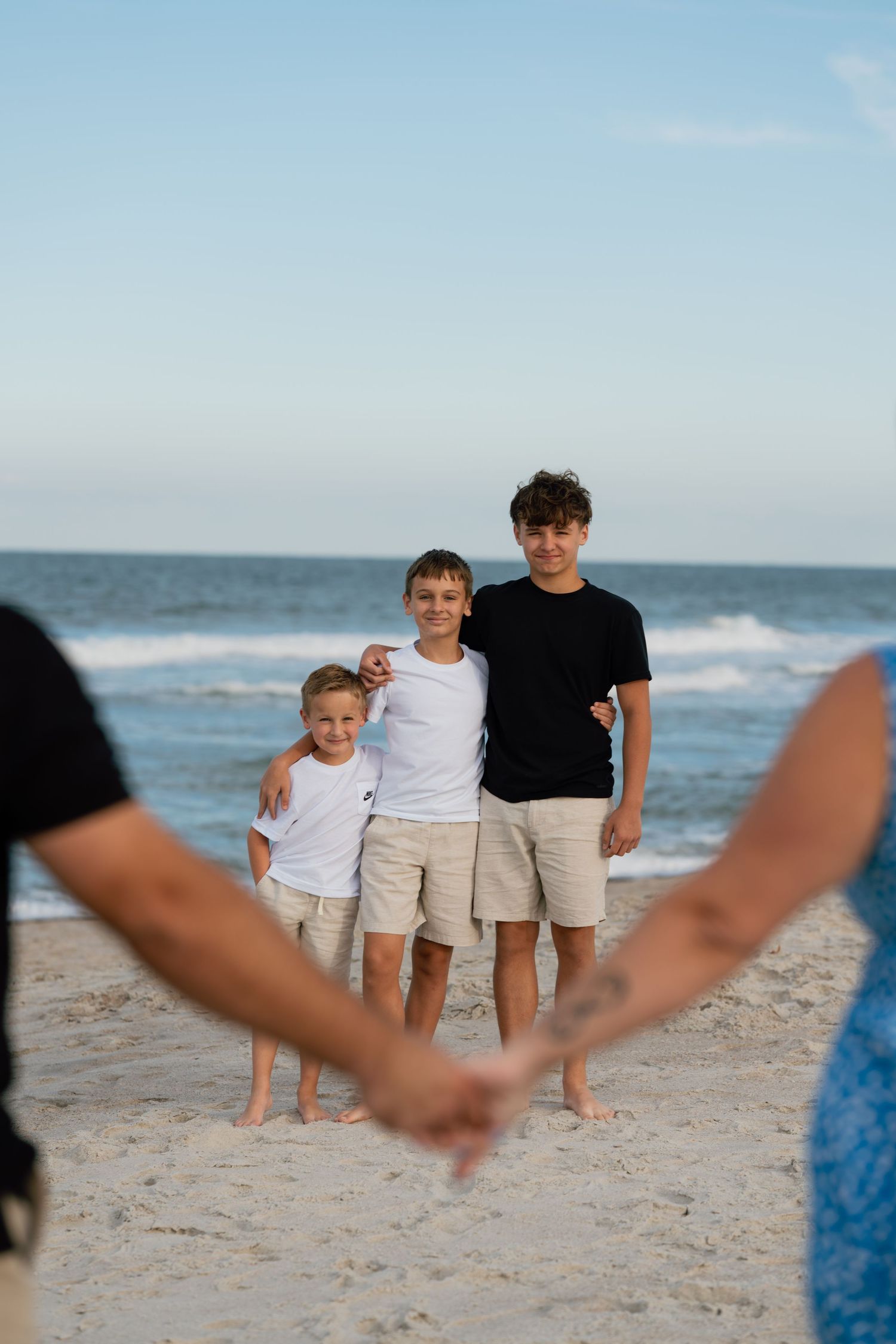 Three boys pose on a sandy beach while two adults hold hands in the foreground framing the scene.