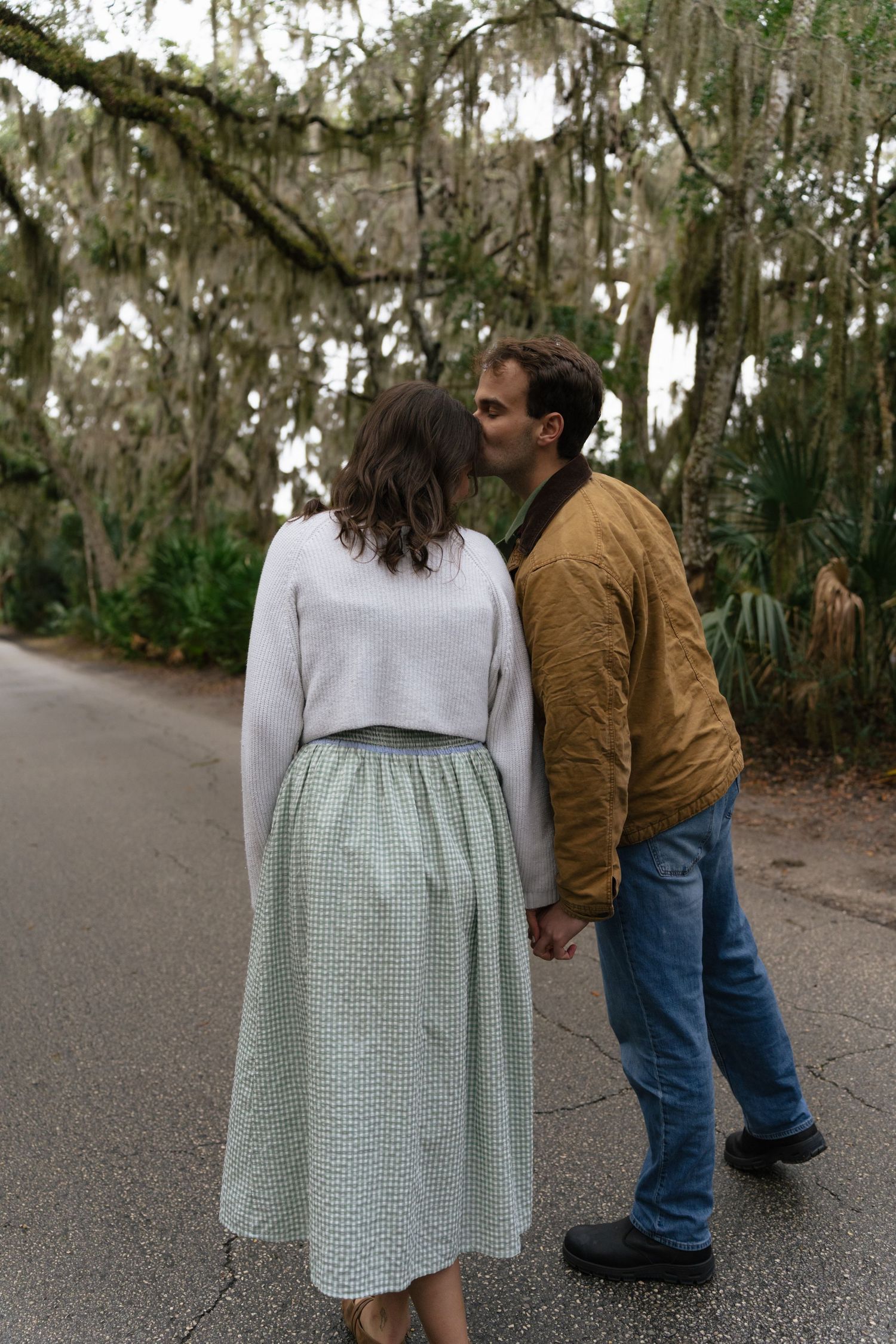 A couple walks hand in hand down a tree-lined path in matching brown and white outfits.