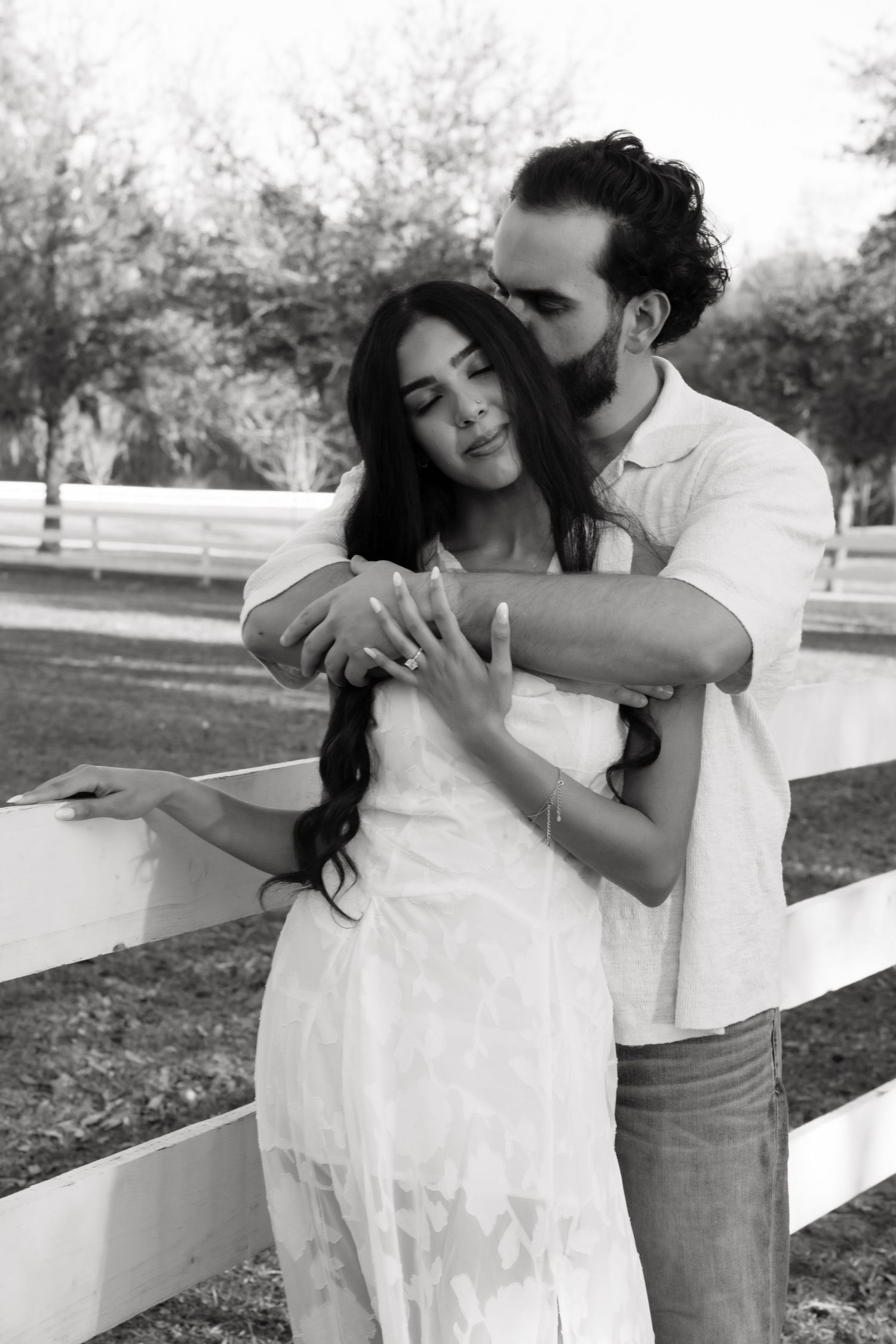 A romantic couple in white clothing embraces lovingly against a white fence in a park setting.