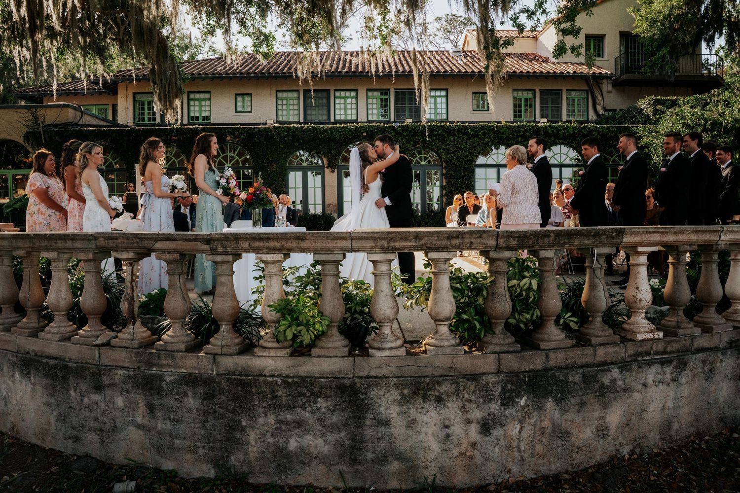 Wedding party stands along stone balustrade at historic venue during outdoor celebration.