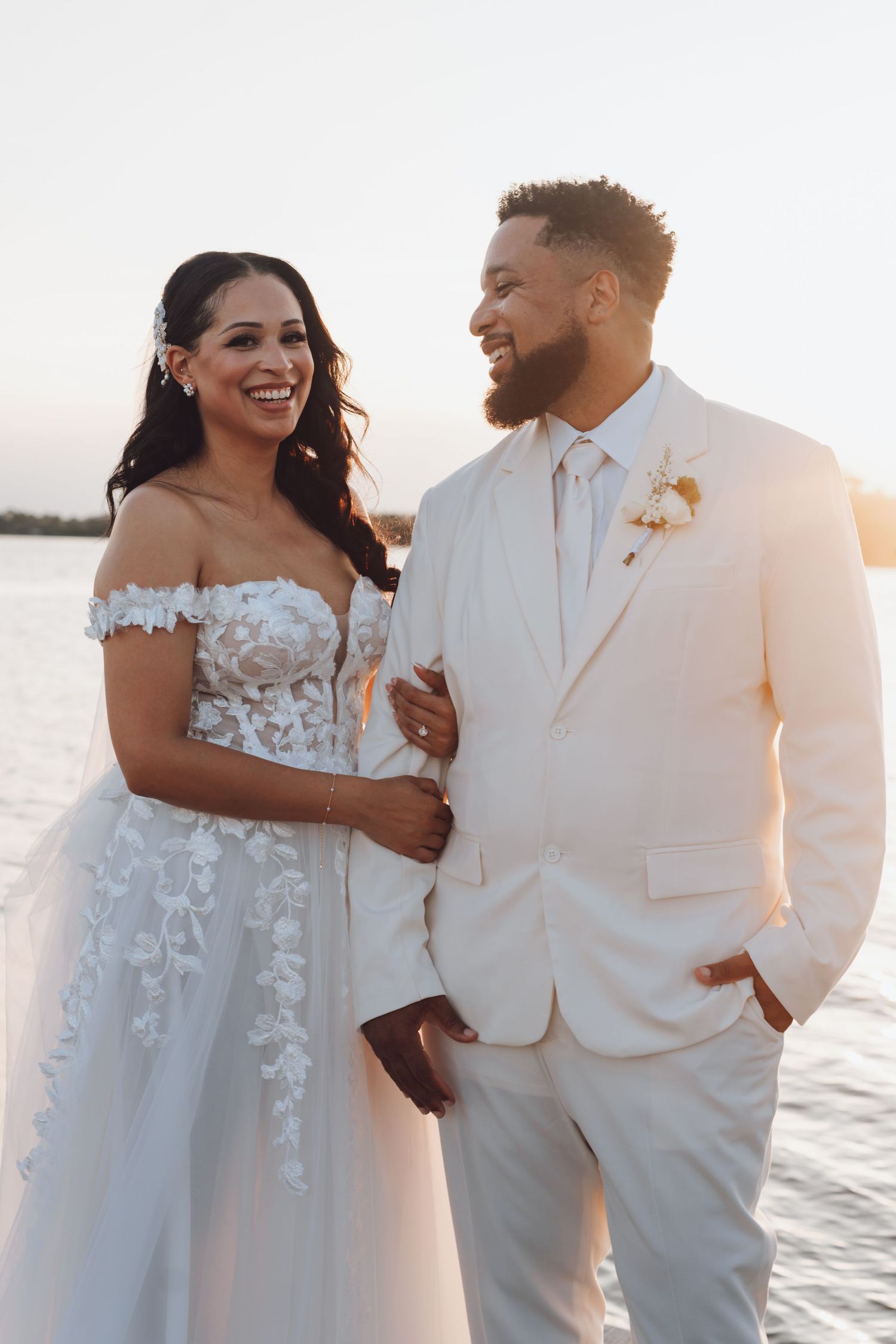 A couple shares a joyful moment at sunset during their beach wedding, wearing white formal attire.