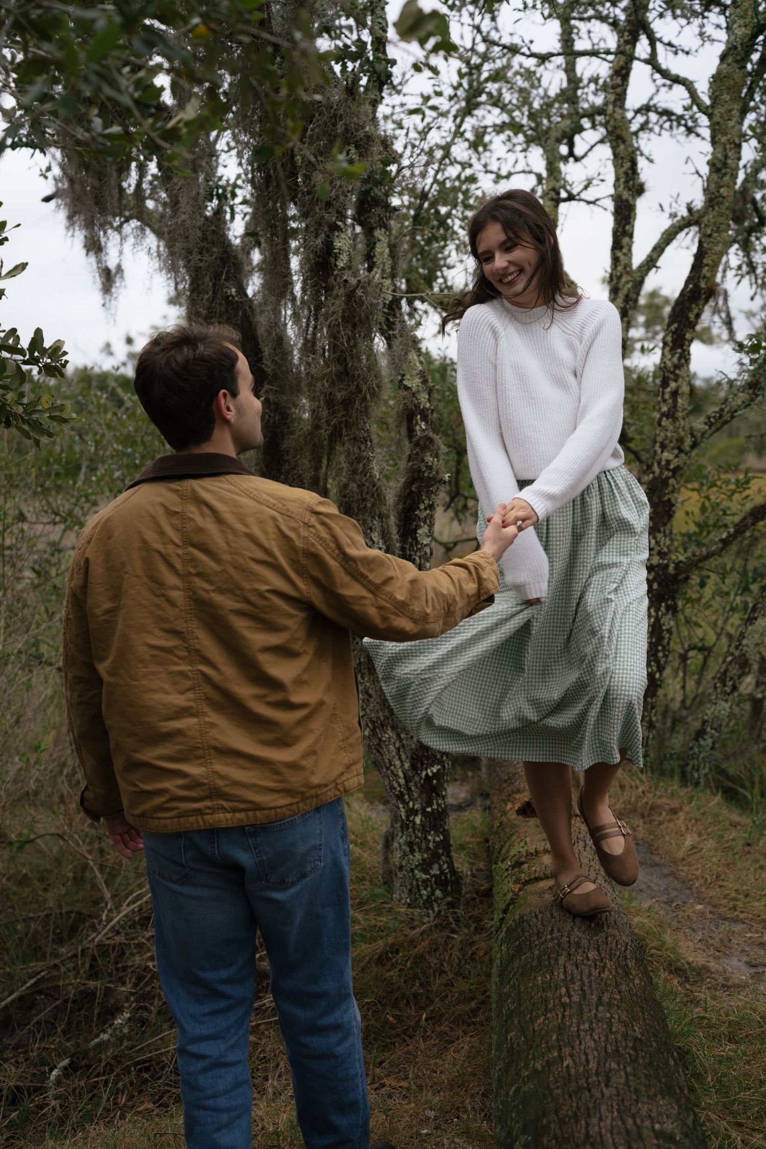 Two people in casual attire interact playfully near trees on a forest path.