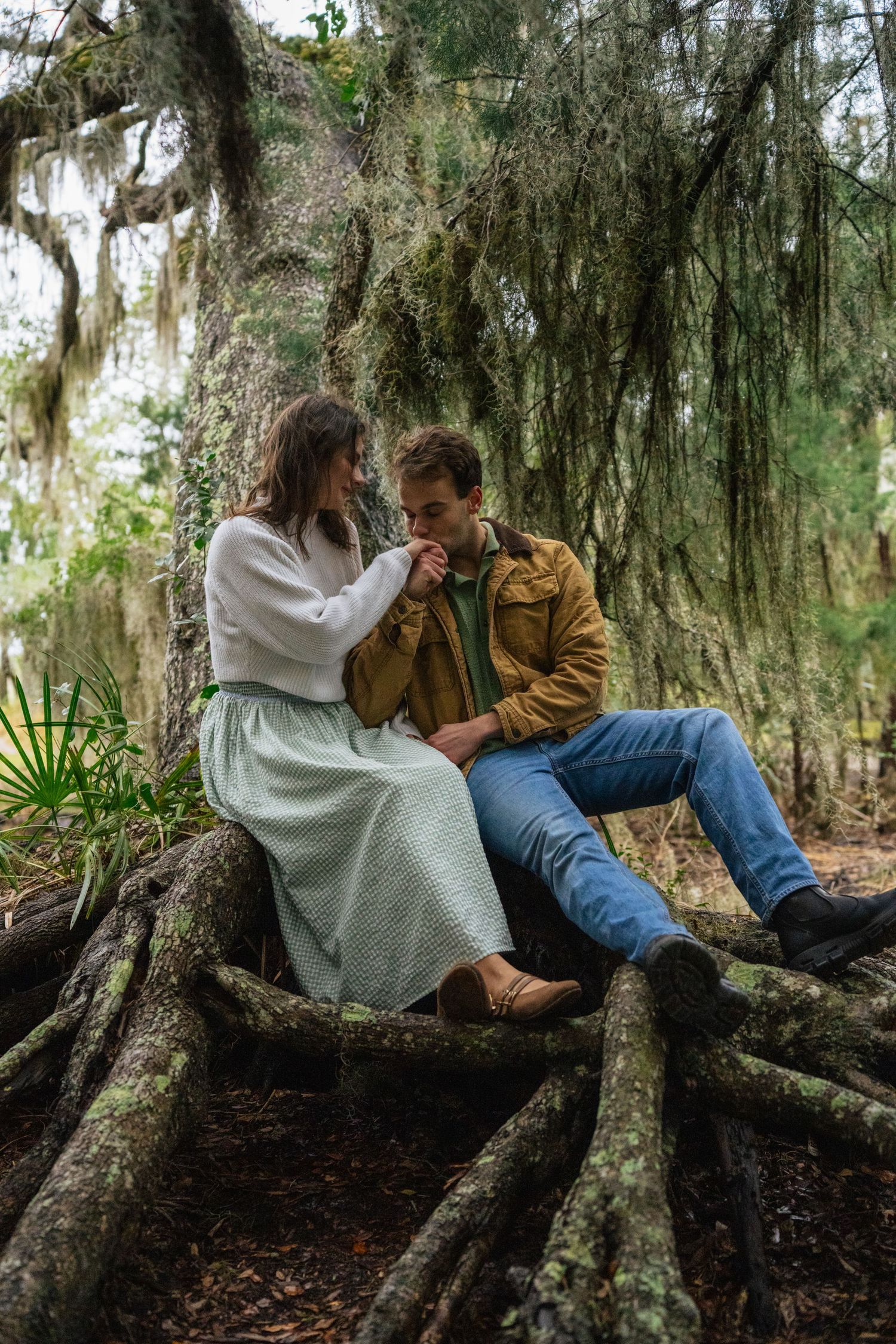 A couple shares an intimate moment while sitting on tree roots in a mossy forest with Spanish moss hanging overhead.
