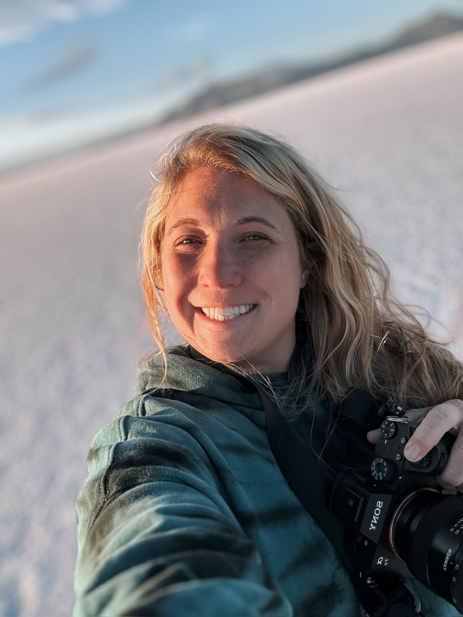 A photographer in a green winter jacket smiles warmly while holding a camera during a snowy outdoor shoot.