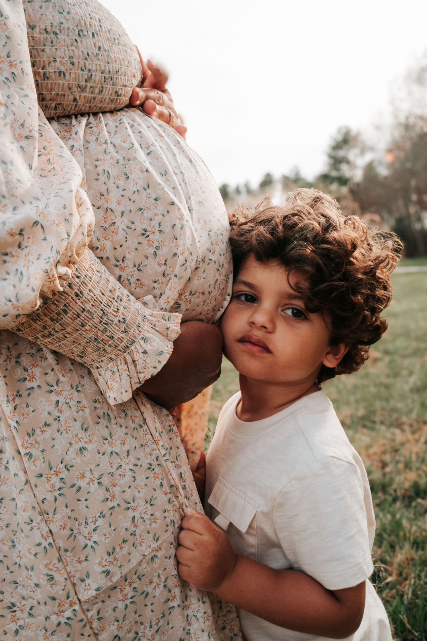 Child in white shirt embraces a floral dress-clad pregnant belly in outdoor setting.