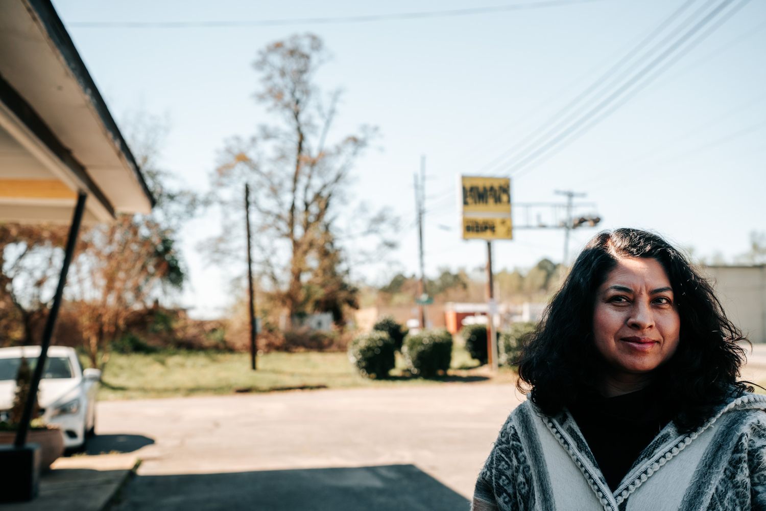 A person stands outside a rural storefront with autumn trees and power lines visible in the background.