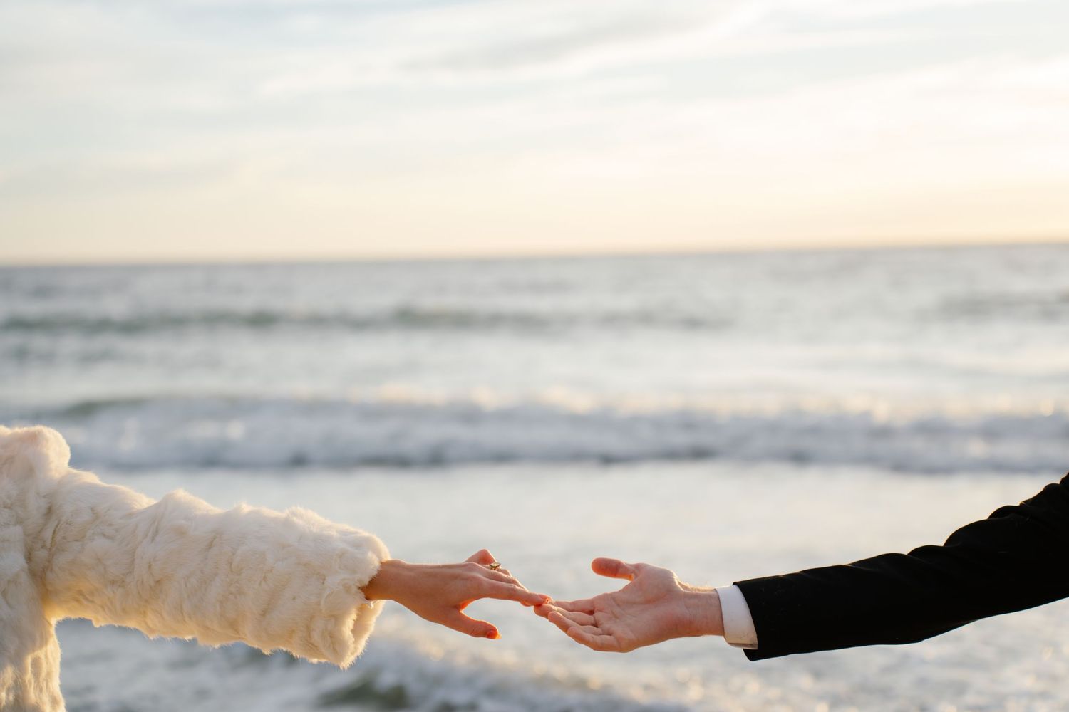 Two hands reaching towards each other against an ocean backdrop with waves and sunset in the background.