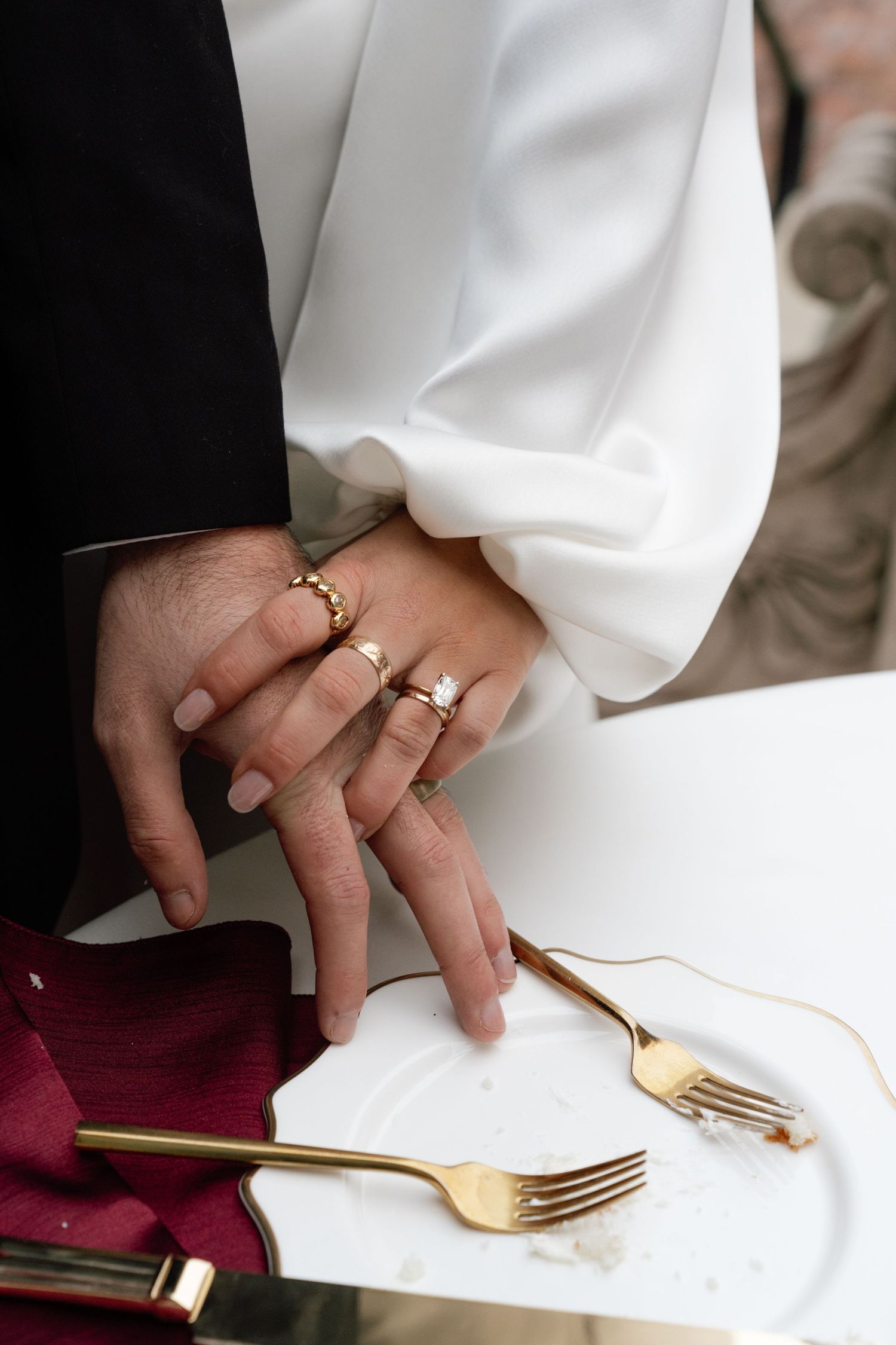 Close-up of newlyweds' hands with wedding rings next to golden forks on white tablecloth with burgundy napkin.