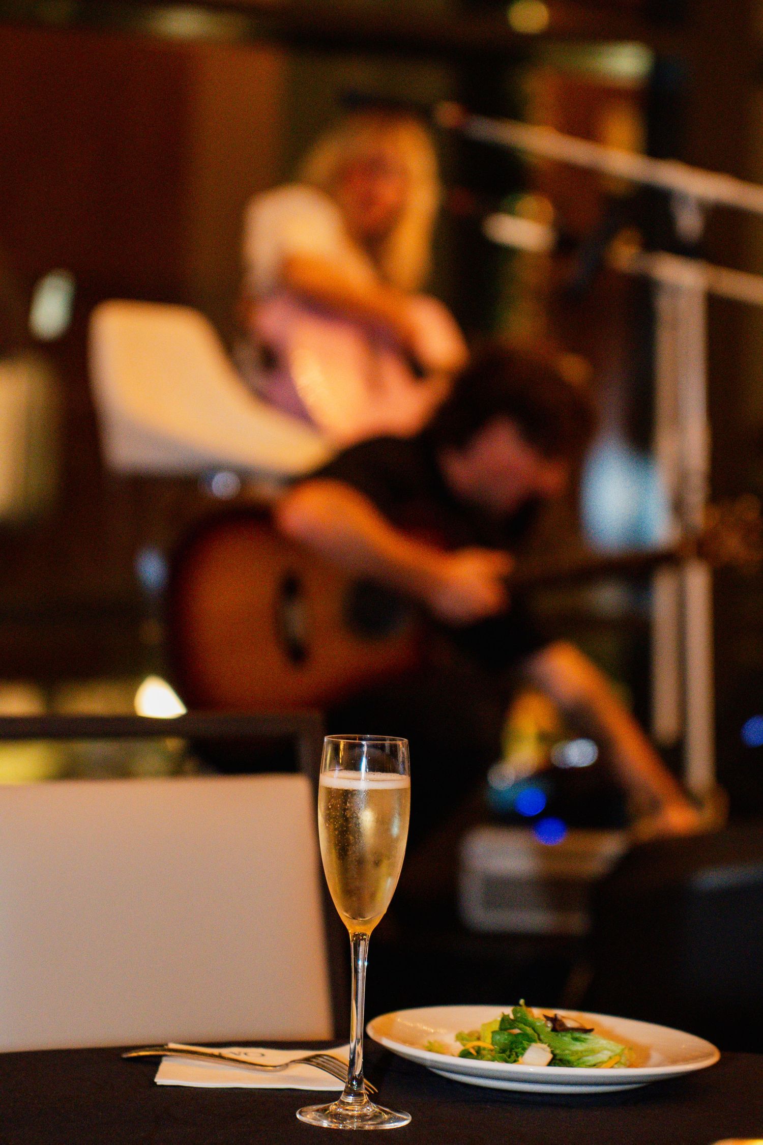 A champagne flute and salad on a table with blurred musicians performing in the background at a restaurant.