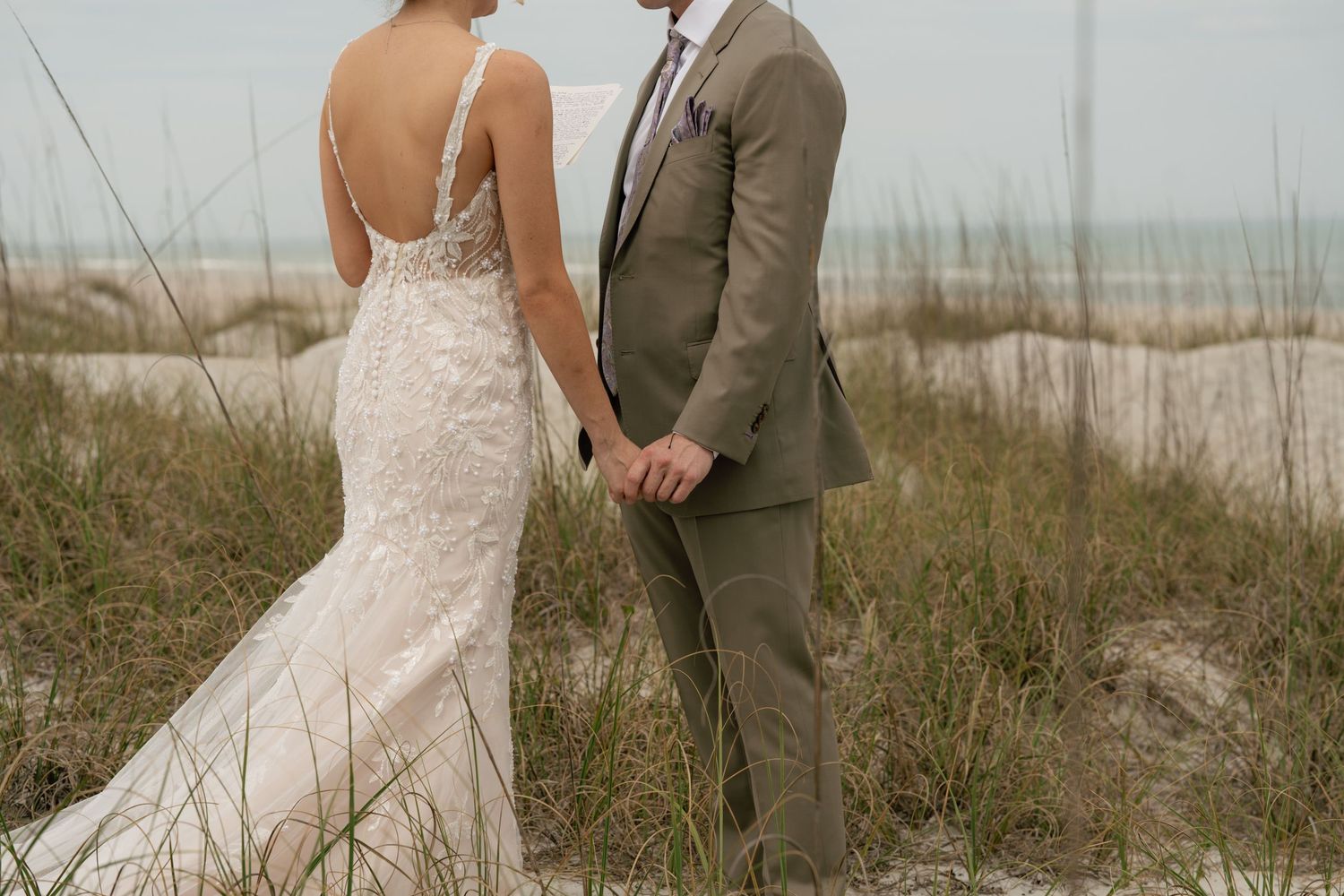 A bride and groom hold hands on a sandy beach, she in a lace mermaid gown, he in a tan suit.