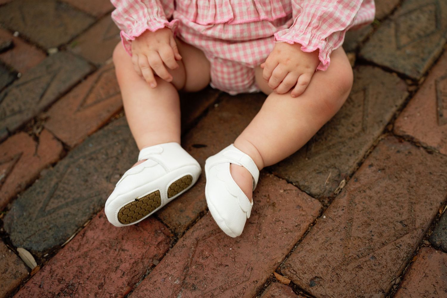 Small white leather shoes and bare legs on a red brick surface with a pink gingham dress visible.