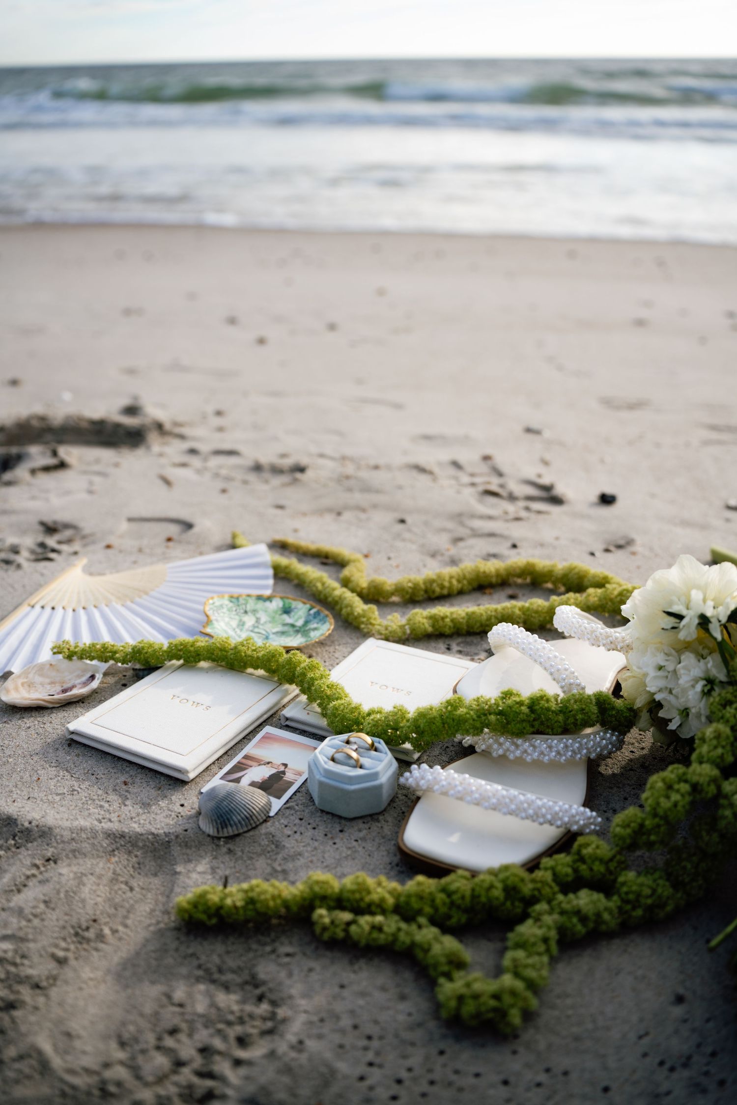 Decorative green moss and white flowers arranged around books and candles on a sandy beach.