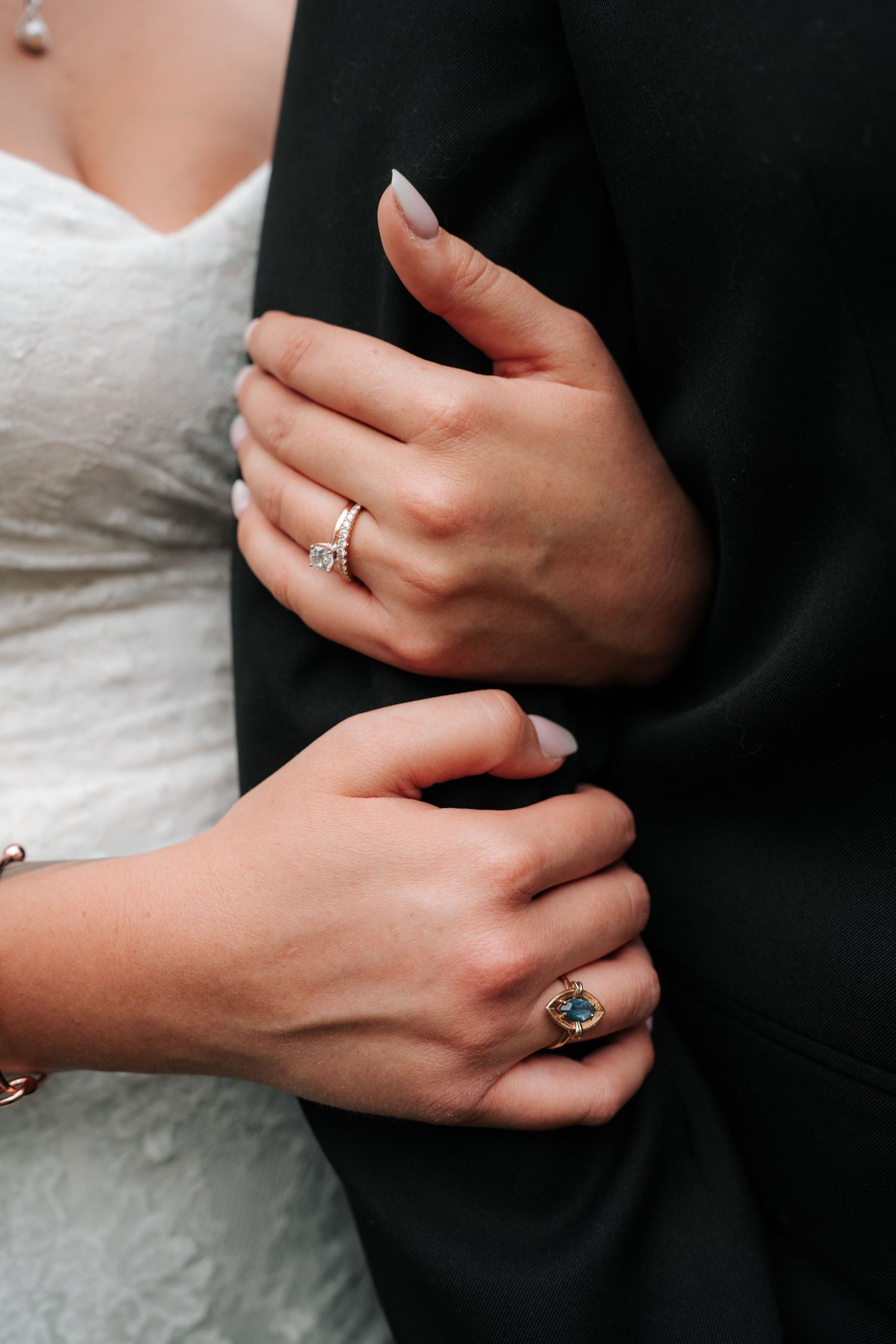 Close-up of elegantly manicured hands with wedding rings embracing against black suit and white dress.