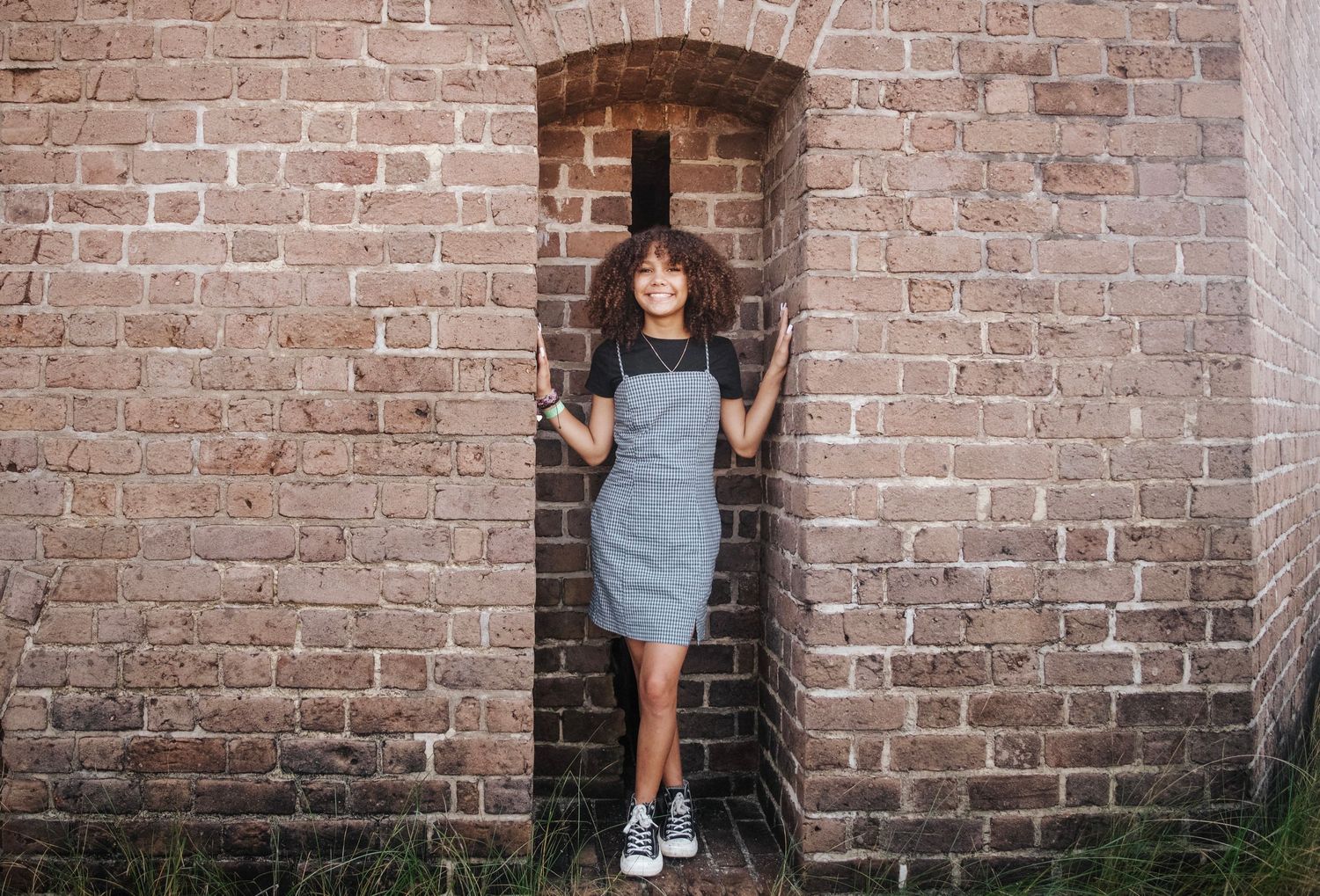 A senior portrait of a girl in a blue and white striped dress and sneakers poses in a rustic brick archway doorway at Fort Clinch State Park