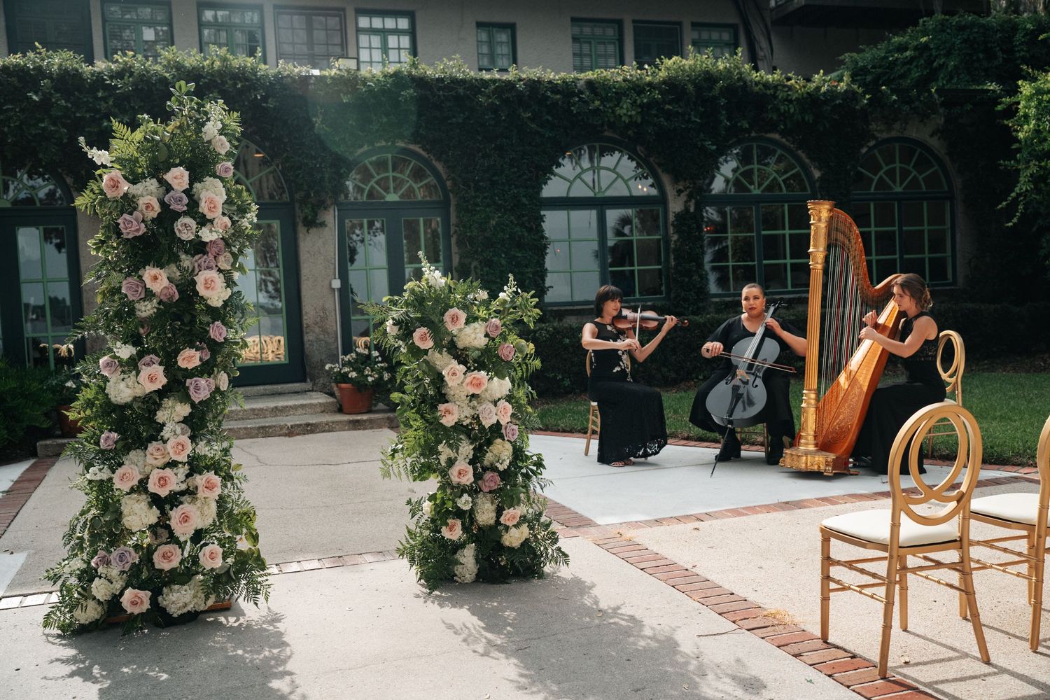 Classical musicians perform with violin and harp beside floral archways in an elegant garden courtyard setting.