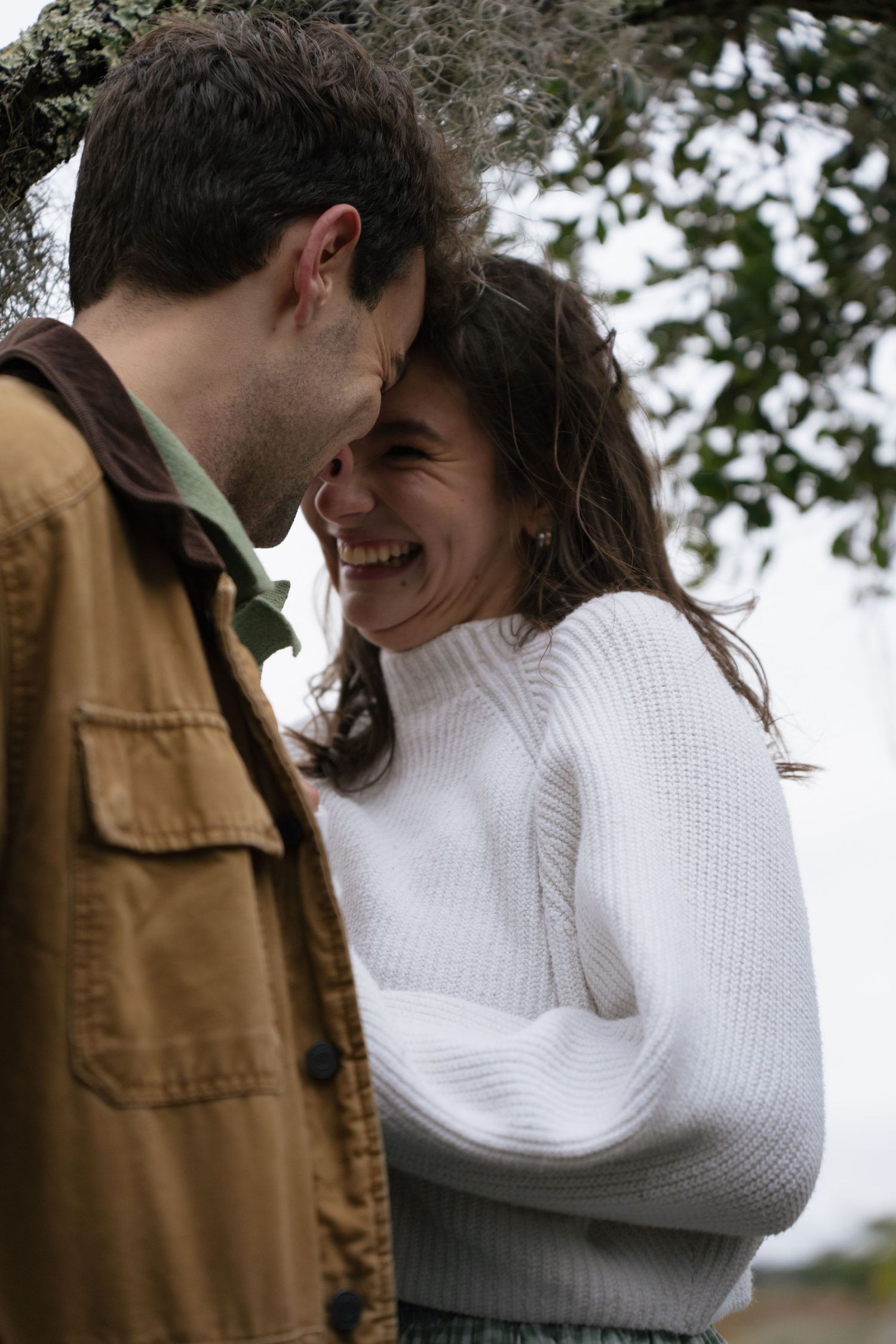 A romantic couple in a white sweater and brown jacket embrace outdoors under trees in a series of intimate moments.