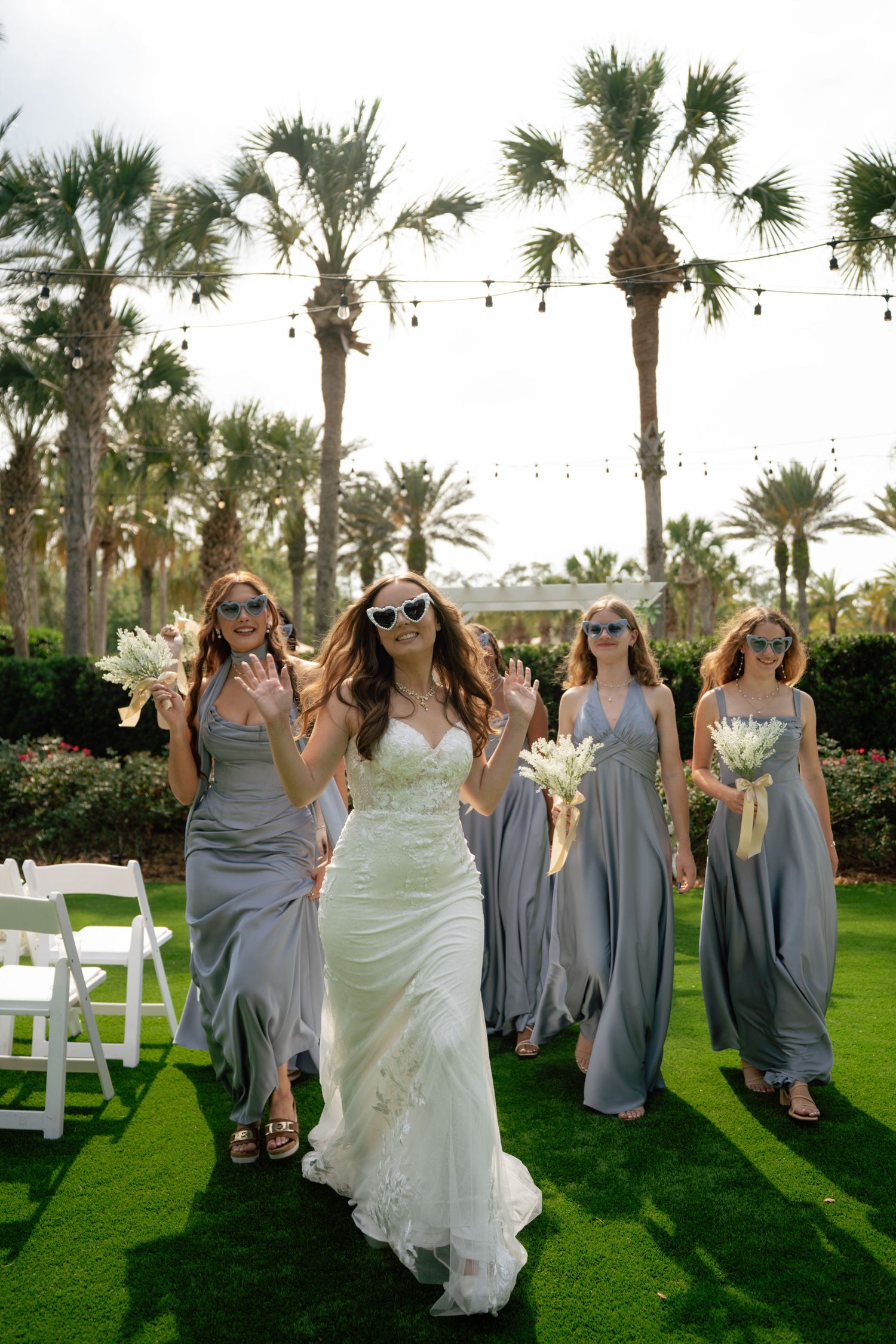 Bride in white gown with bridesmaids in gray dresses waving and holding bouquets on green lawn with palm trees.