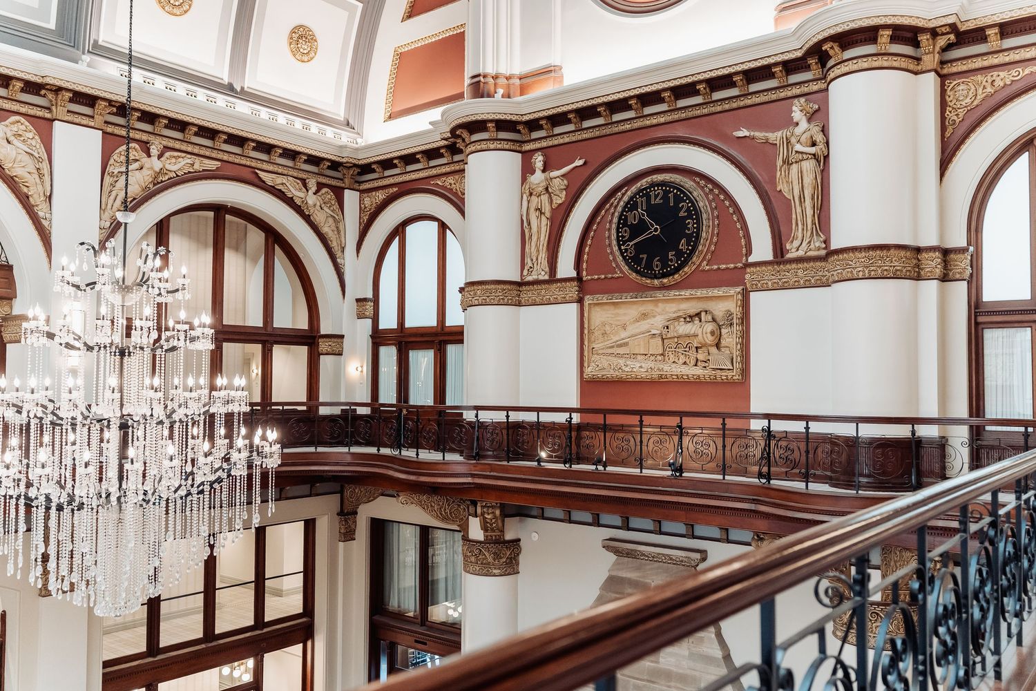 Elegant Victorian interior with crystal chandelier, arched windows, and ornate wooden staircase featuring decorative railings.