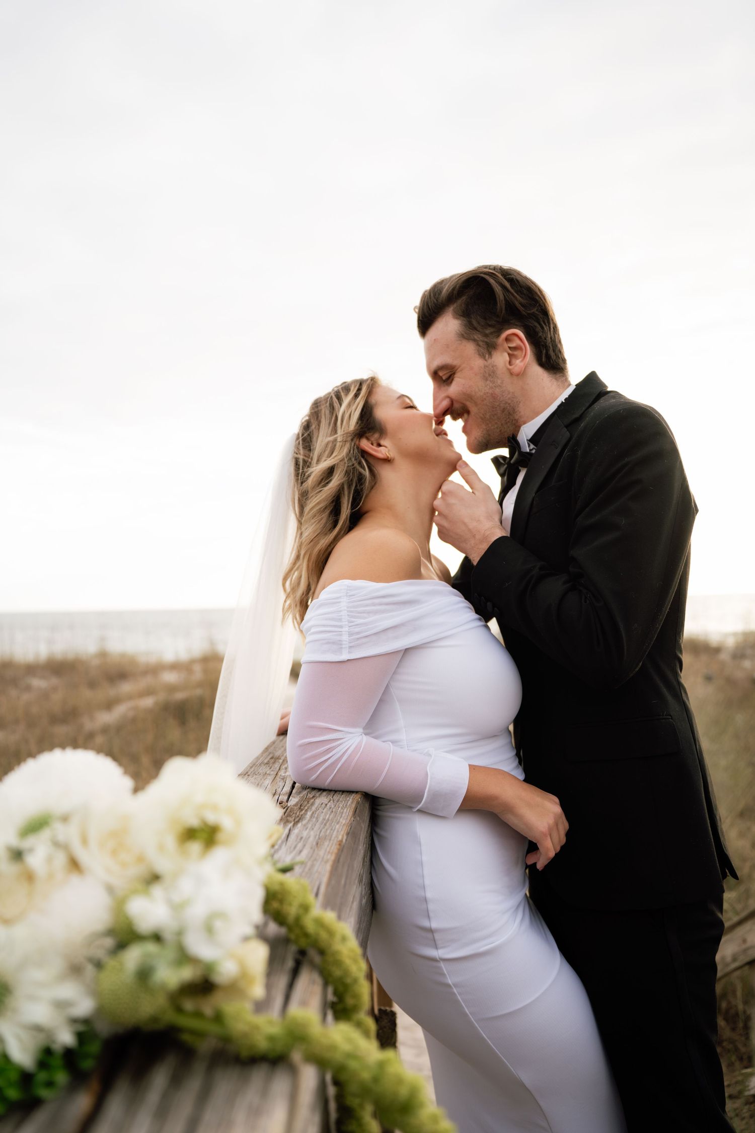 Romantic wedding couple embraces outdoors with white floral arrangements against an overcast sky.