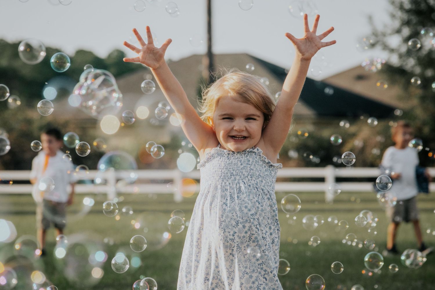 Small figure in white dress throws hands up joyfully amid floating soap bubbles in sunny outdoor setting.