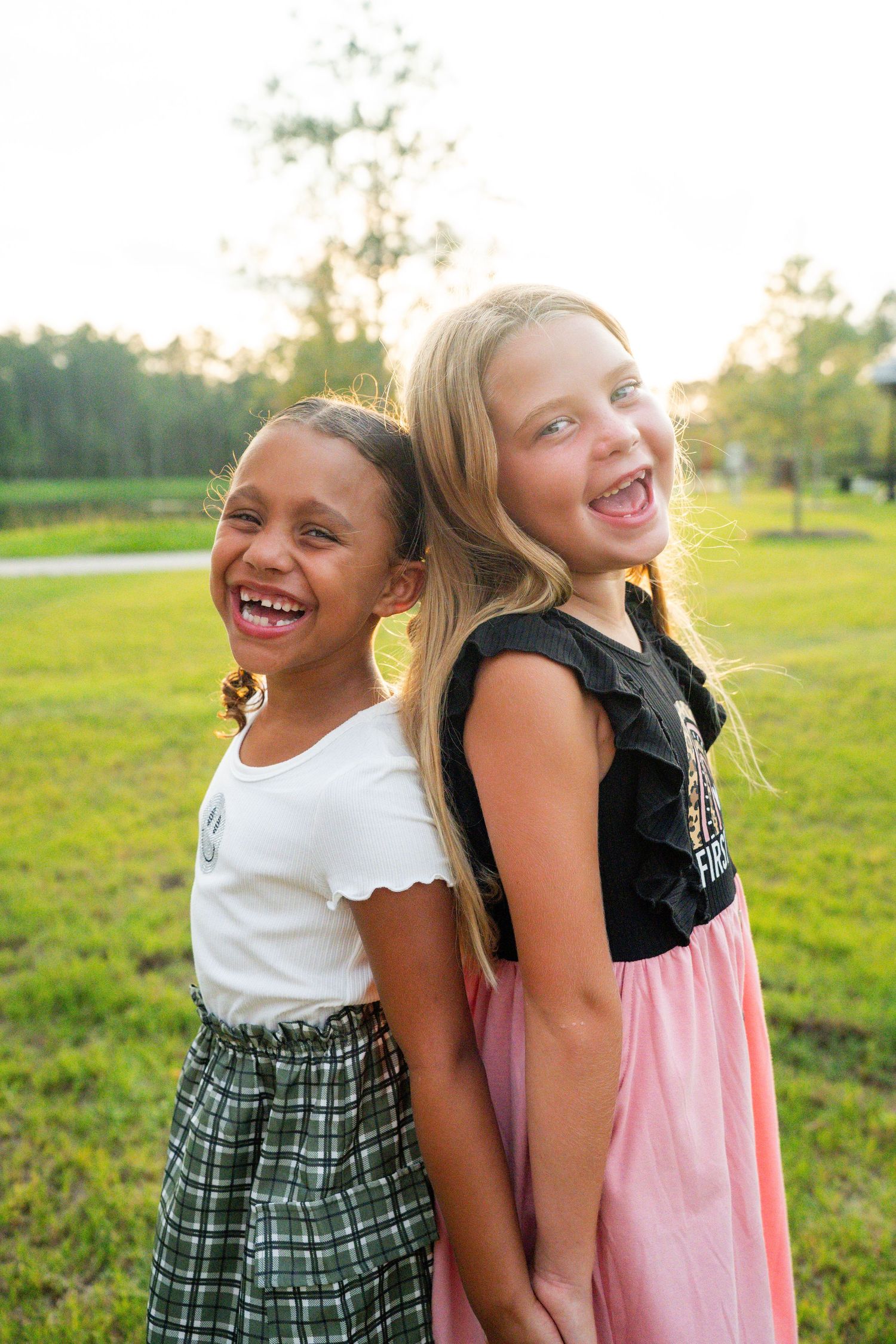 Two children laughing together while standing back-to-back in a sunny grassy field at sunset.