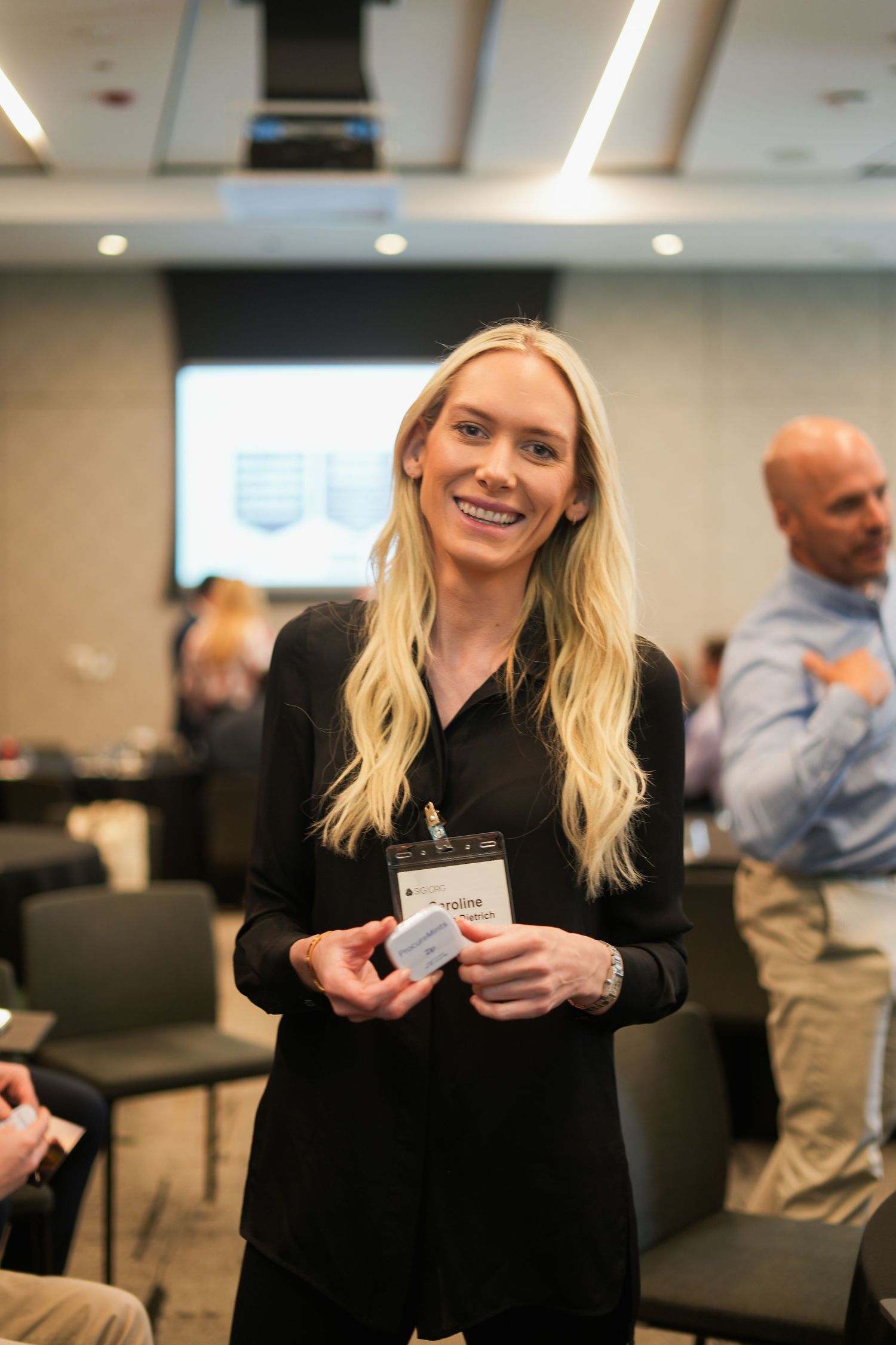 Smiling blonde woman in black outfit wearing conference name badge at professional networking event.