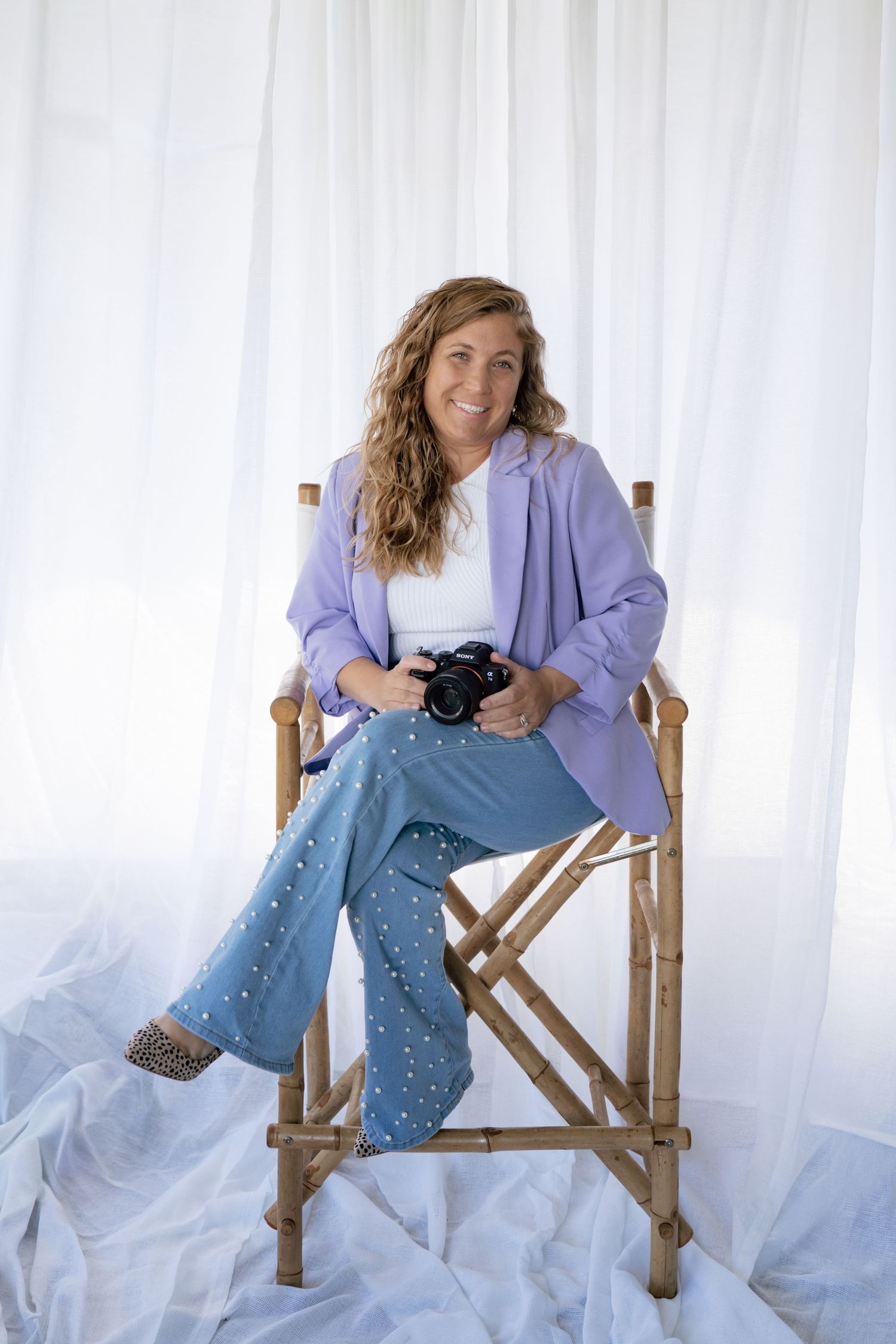 A photographer in a purple blazer and jeans poses casually in a wooden director's chair against a white backdrop.