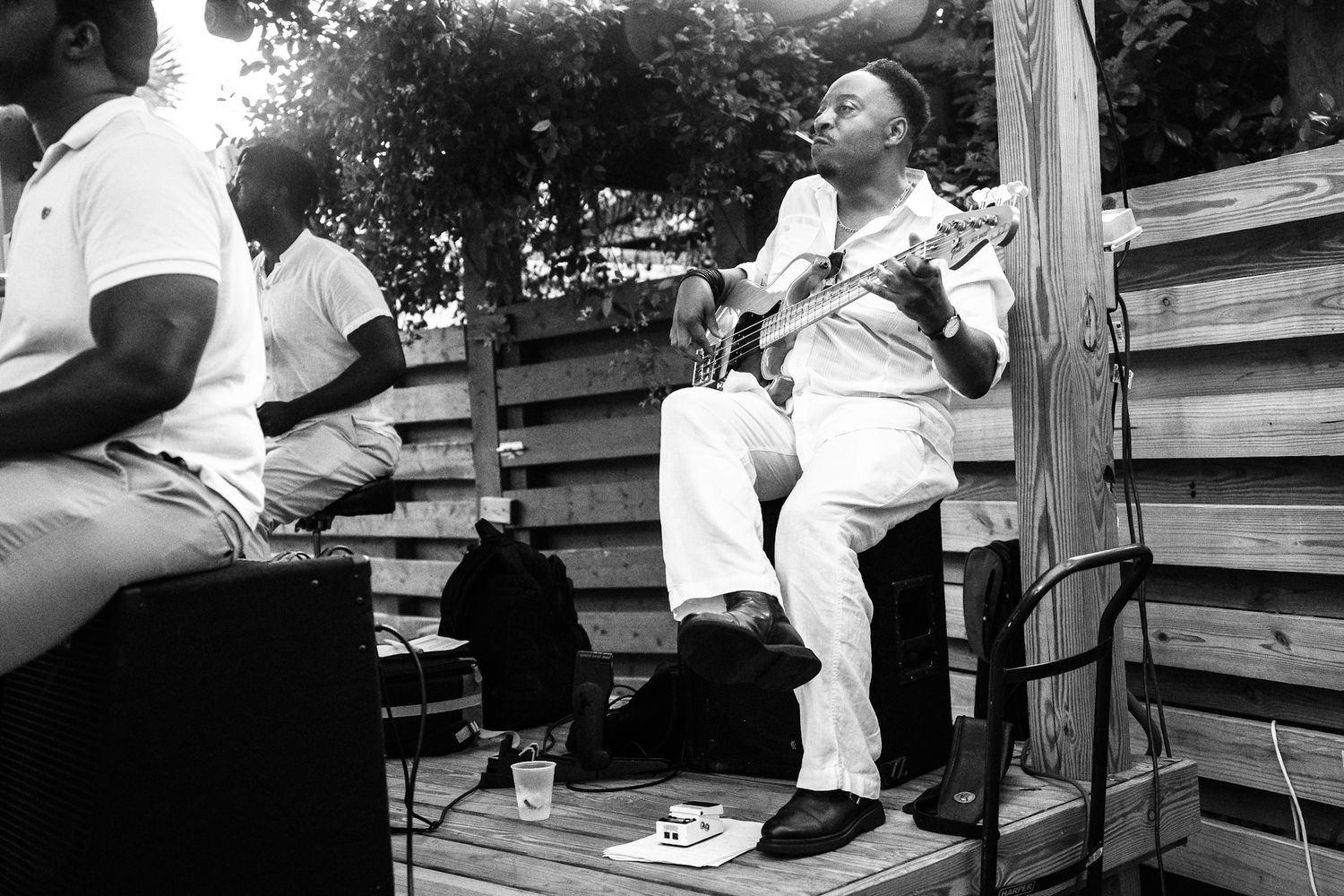 Black and white photo of a musician playing guitar on an outdoor wooden stage while seated, performing for a small audience.